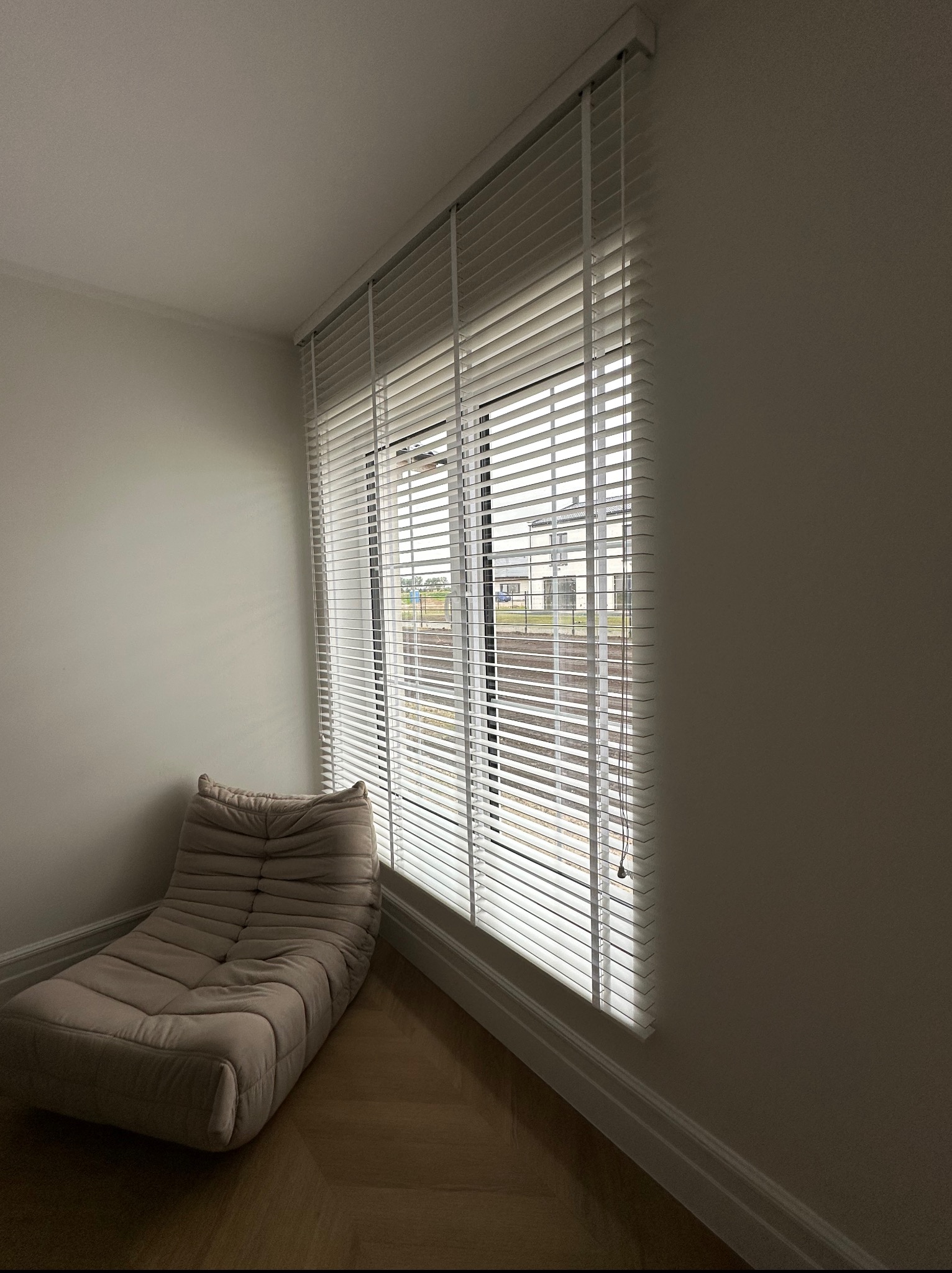 Jasne, drewniane żaluzje poziome zamontowane w oknie z widokiem na dom. Obok okna beżowy, miękki fotel na drewnianej podłodze. Stonowana kolorystyka wnętrza.