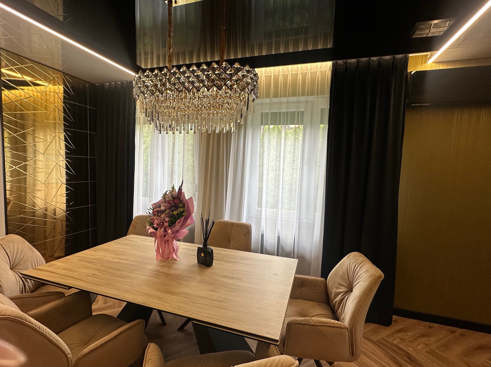 Elegancka jadalnia z beżowymi krzesłami, drewnianym stołem i kryształowym żyrandolem na tle okna z zasłonami. Wazon z kwiatami na stole.
