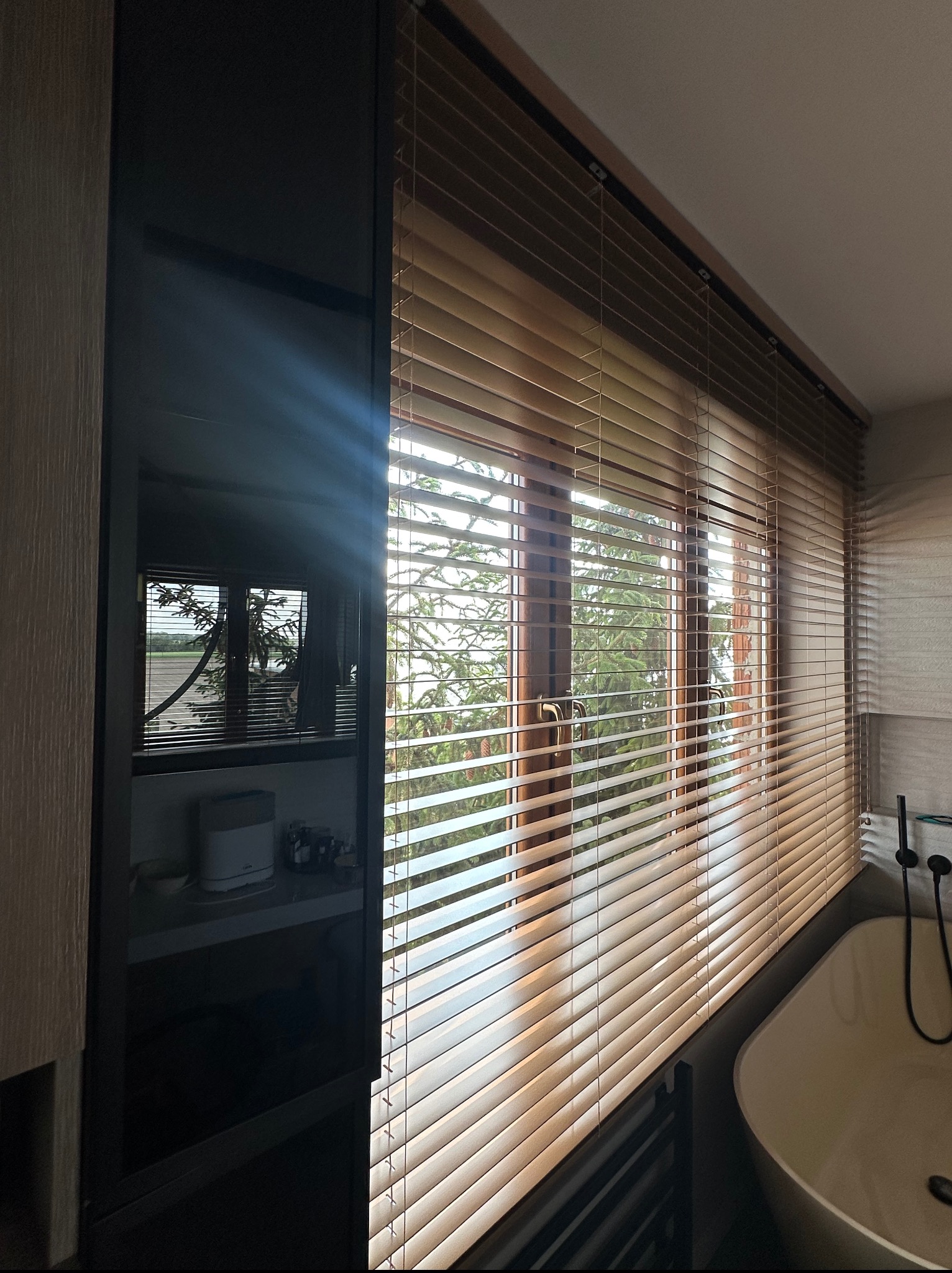Jasne, drewniane żaluzje poziome w łazience, z widokiem na zieleń za oknem. Minimalistyczny design, ciepłe oświetlenie.