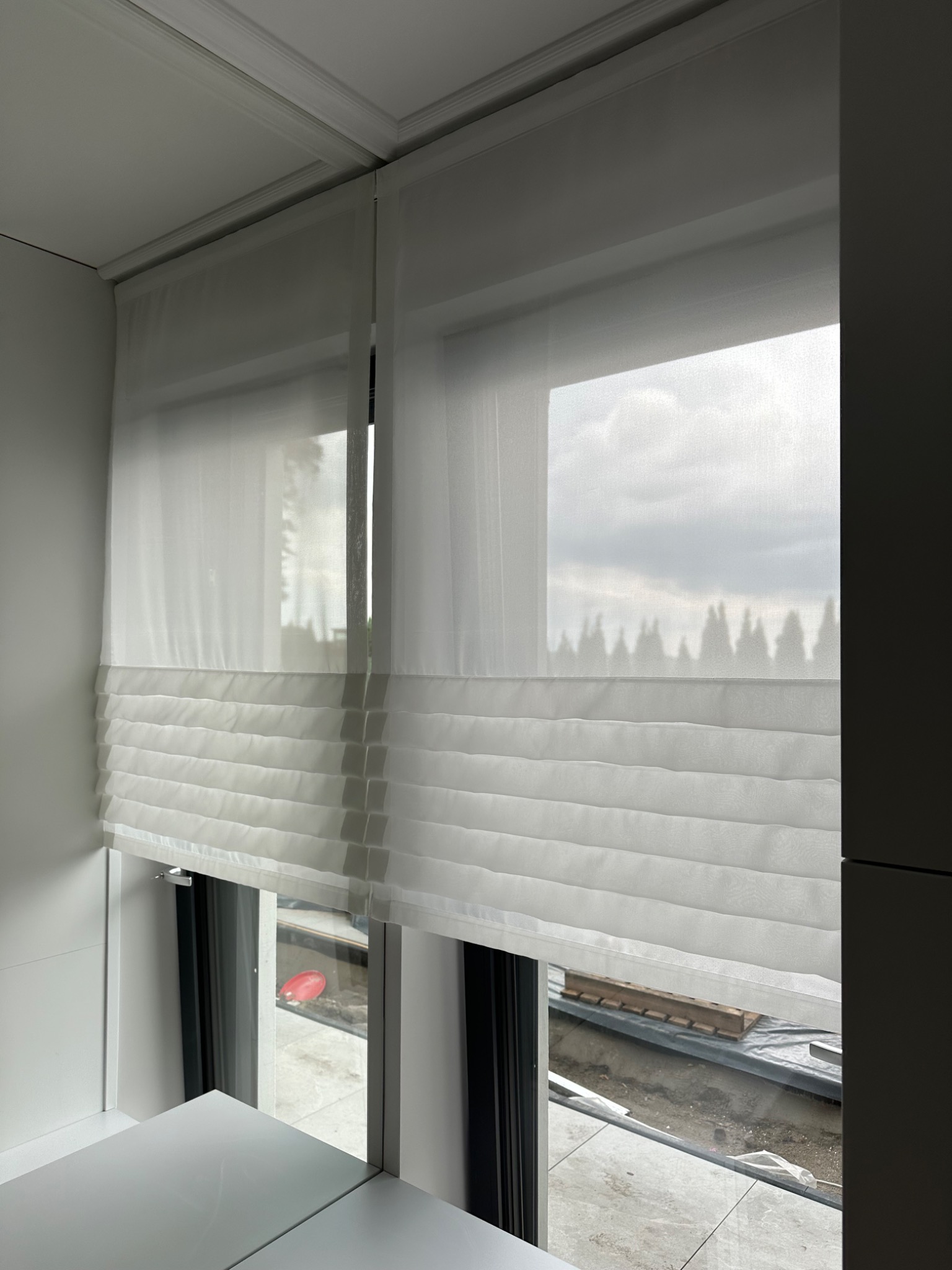 Delikatna, biała roleta rzymska z poziomymi fałdami, zamontowana w oknie. Za oknem widoczne drzewa i pochmurne niebo.