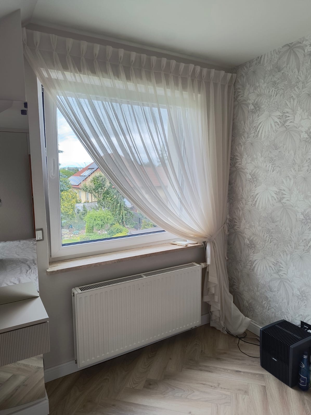 Jasna, zwiewna zasłona upięta sznurem przy oknie z widokiem na zieleń, na tle ściany w roślinny wzór. Pod oknem biały grzejnik.