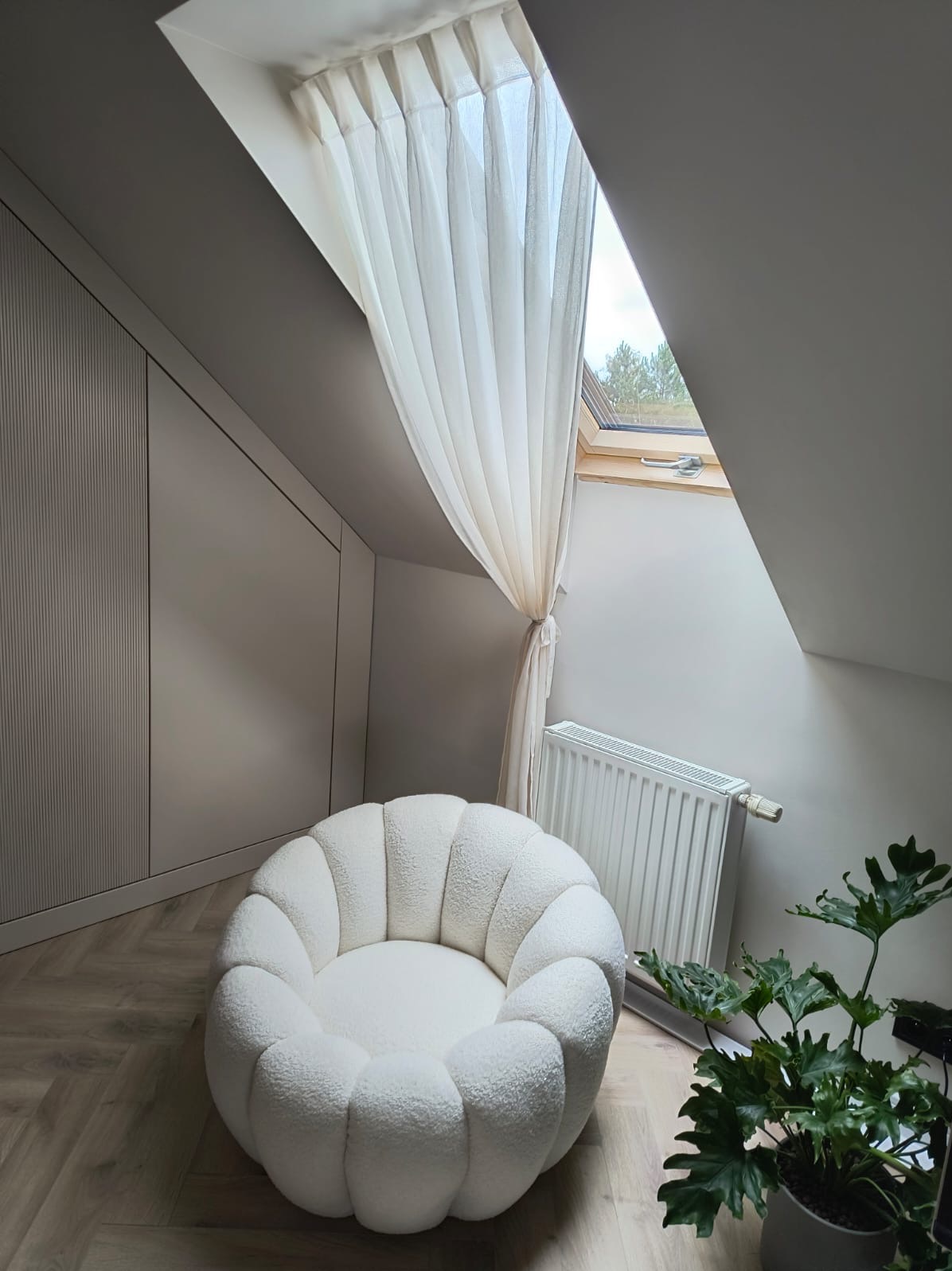 Jasna, zwiewna firanka przy oknie dachowym w skosie poddasza, obok dekoracyjny fotel i grzejnik. Minimalistyczne wnętrze w odcieniach bieli i szarości.