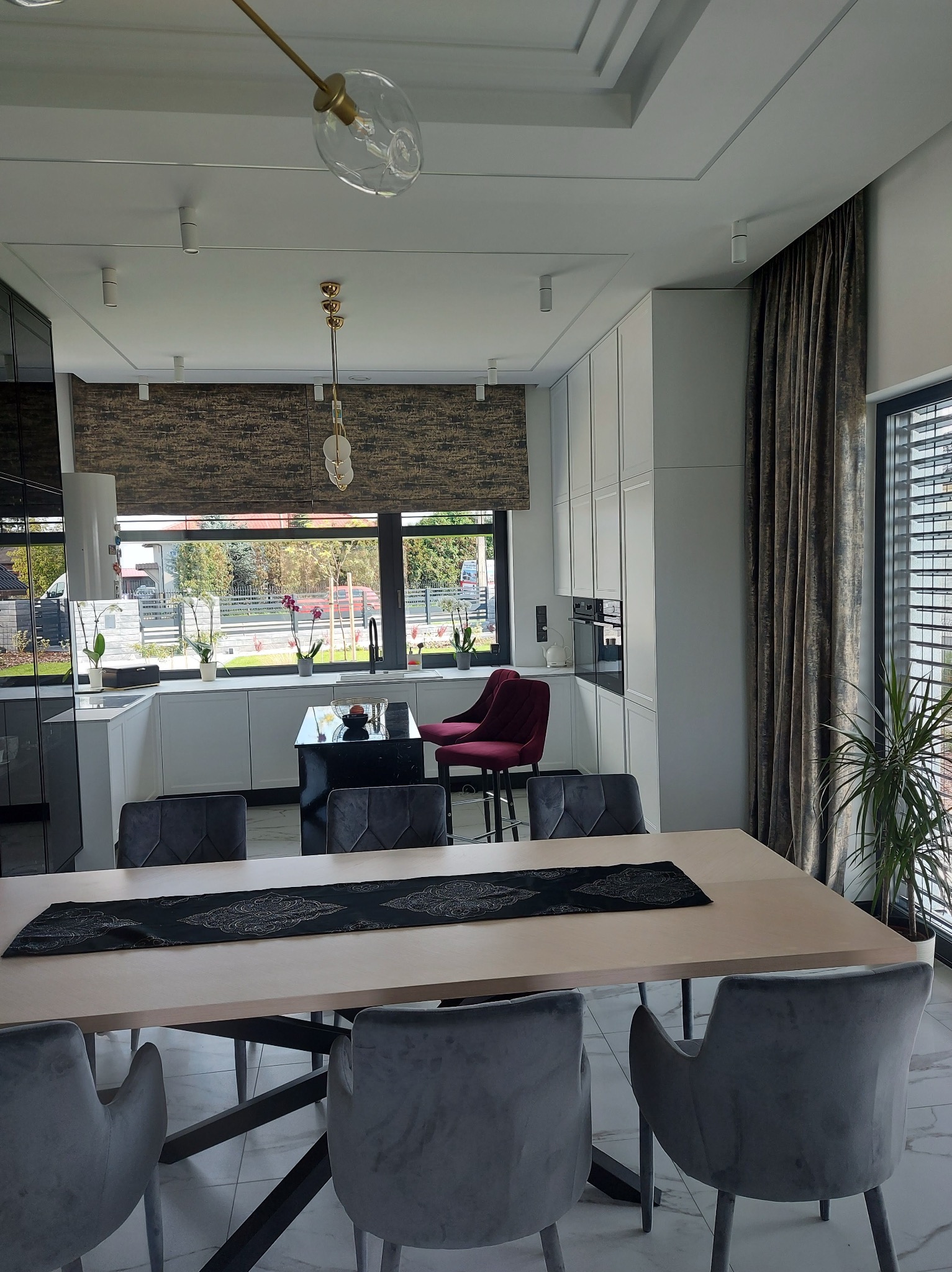 Eleganckie wnętrze: stół z szarymi krzesłami, w tle kuchnia z roletą w oknie i zasłoną przy oknie. Minimalistyczny design z akcentem roślinnym.