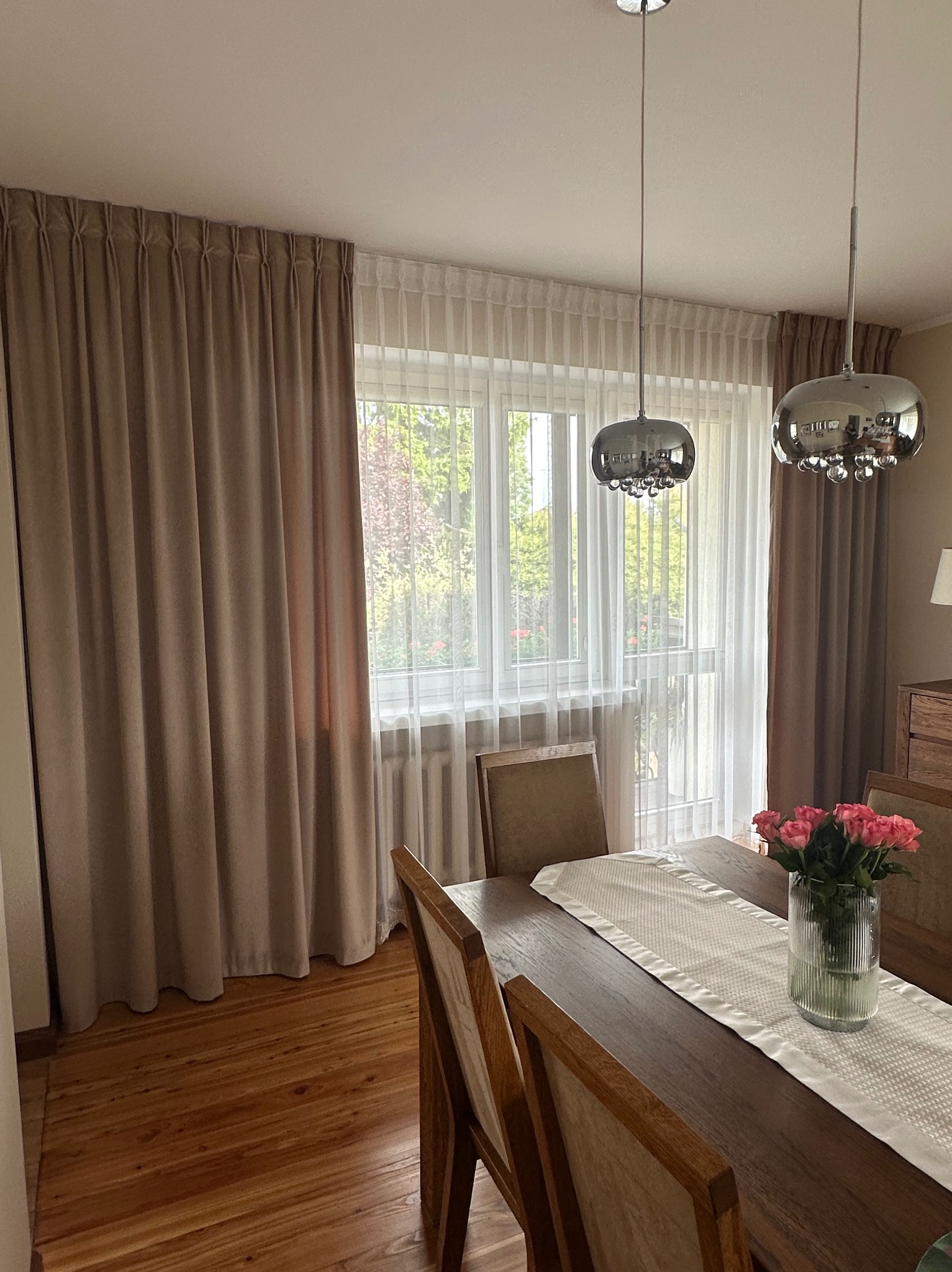 Beżowe zasłony i firany w oknie jadalni, drewniany stół z bieżnikiem i wazonem róż, nowoczesne lampy wiszące. Widok na ogród przez okno.