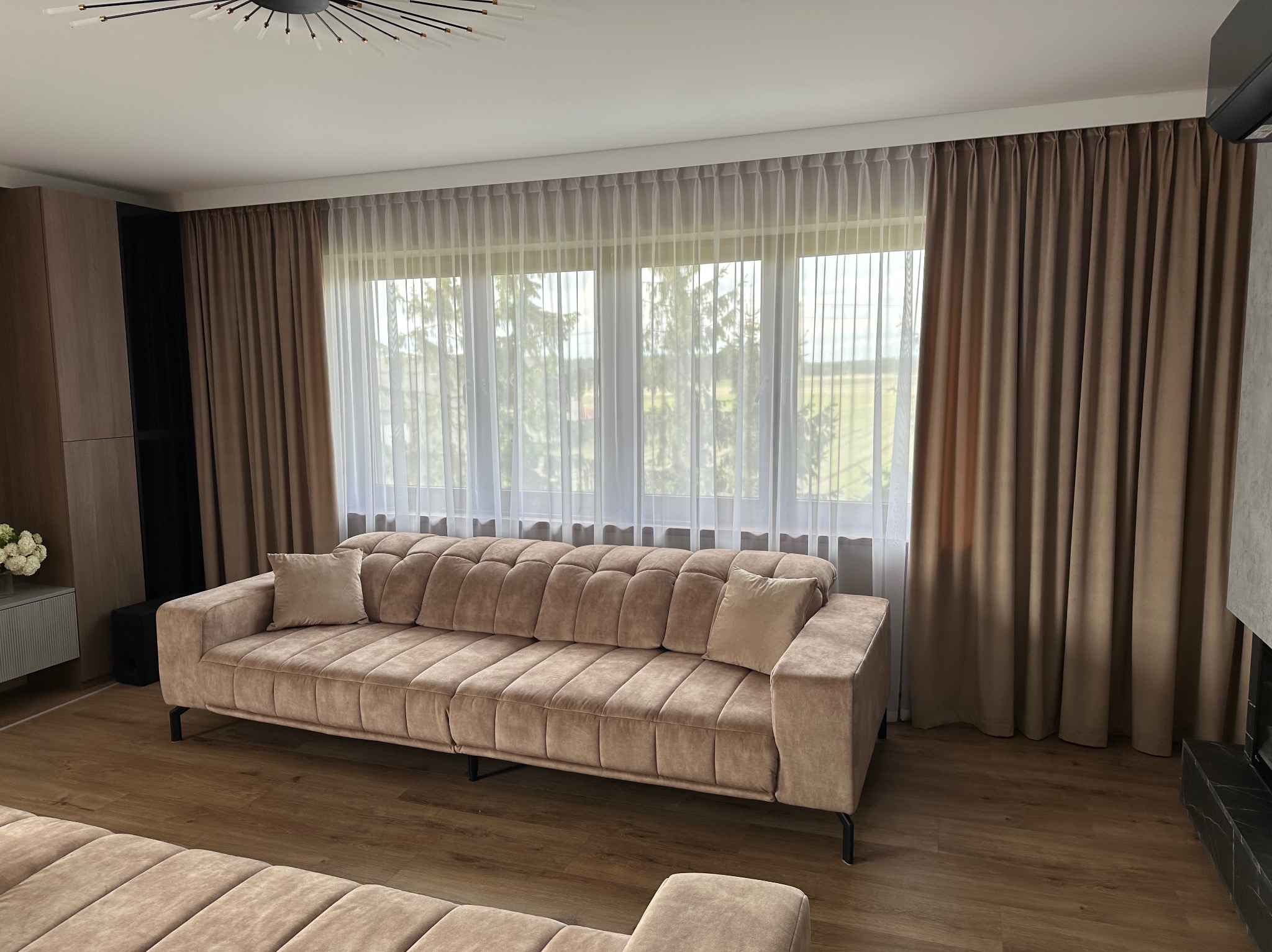 Beżowe zasłony i firany w salonie z sofą w podobnym odcieniu. Widok na zewnątrz przez okno. Subtelne światło dzienne.