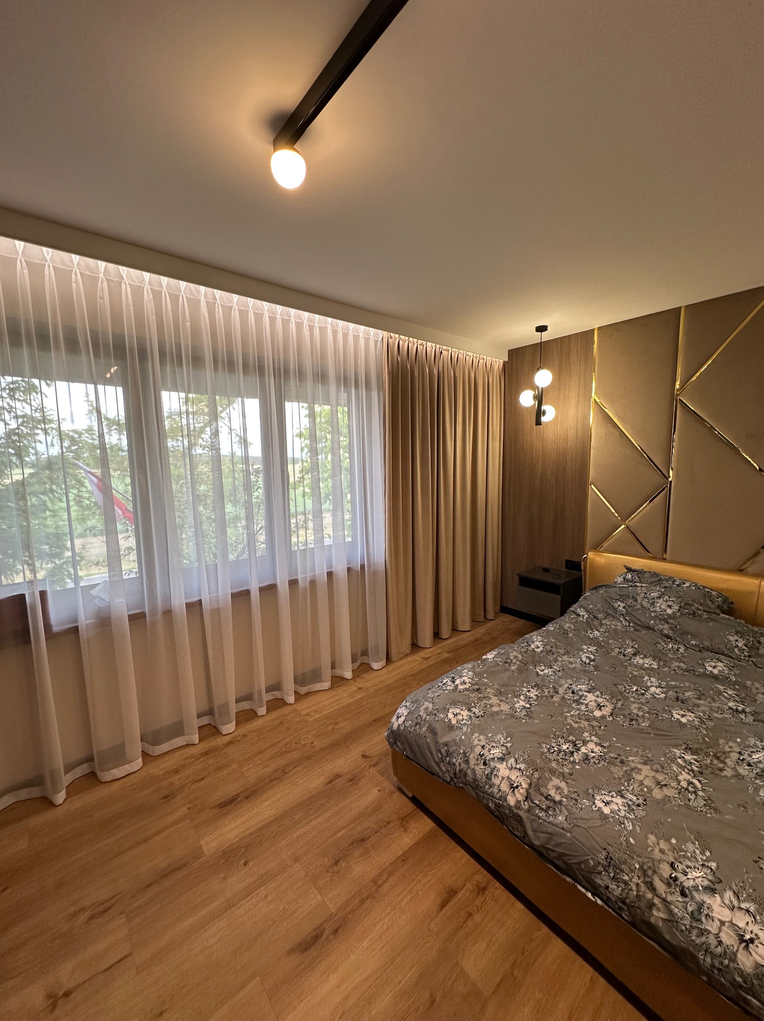 Elegancka sypialnia z oknem przesłoniętym zwiewnymi, białymi zasłonami i beżowymi zasłonami zaciemniającymi. Dekoracyjna ściana za łóżkiem.