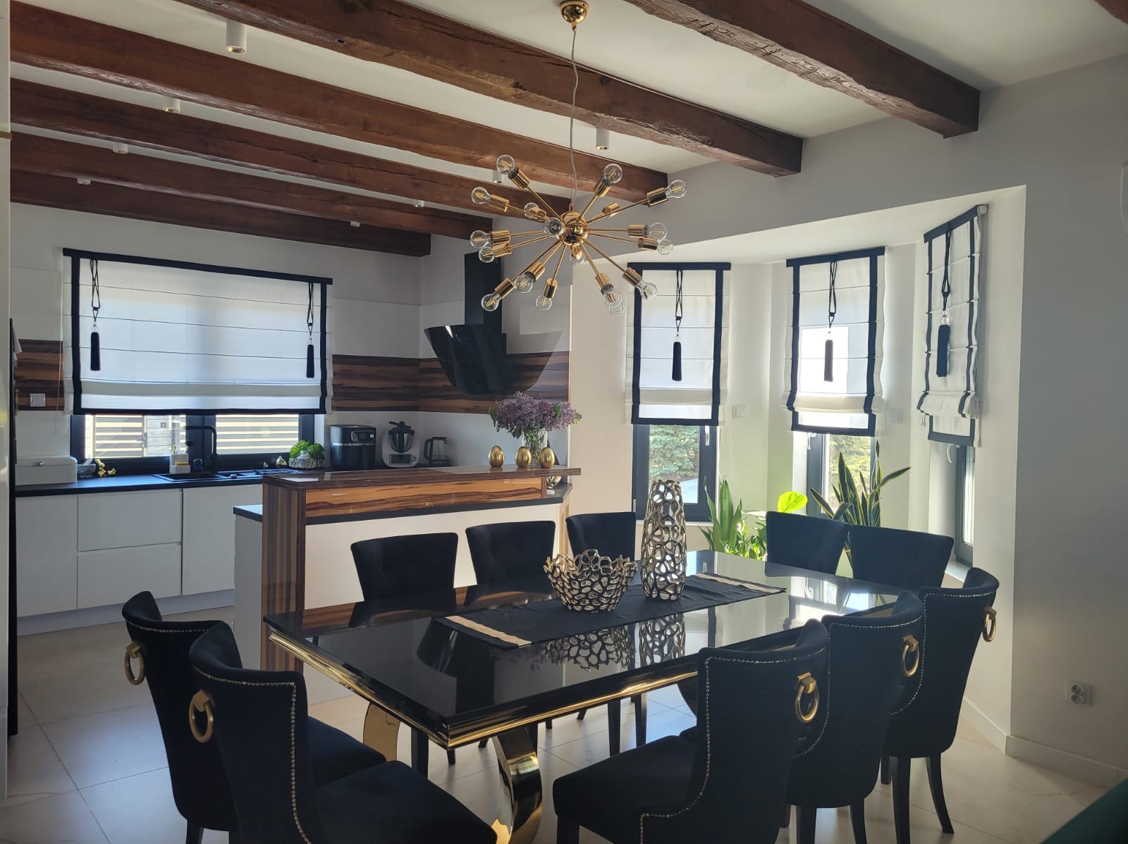 Eleganckie rolety rzymskie w jasnym wnętrzu jadalni z czarnymi krzesłami i złotymi akcentami. Widoczne drewniane belki sufitowe i nowoczesna lampa.