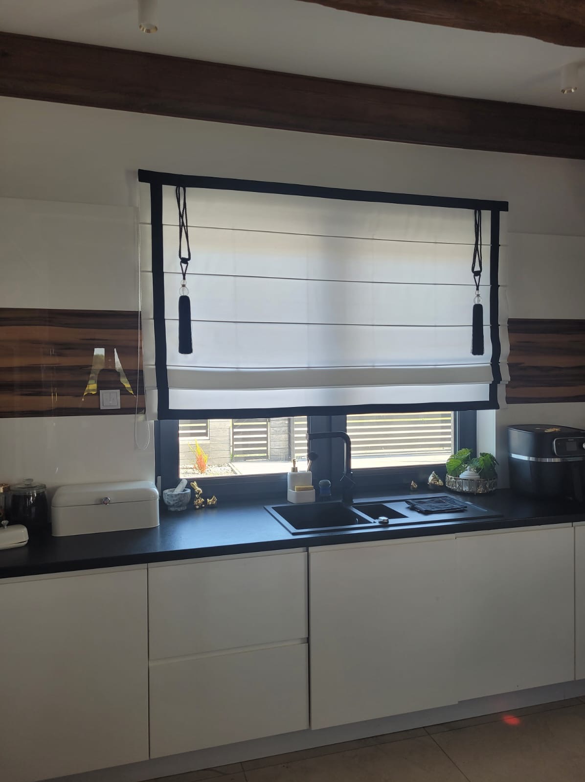 Elegancka, biała roleta rzymska z czarnym wykończeniem w kuchni, zawieszona nad oknem z widokiem na zewnątrz. Minimalistyczny design, drewniany sufit i białe szafki.