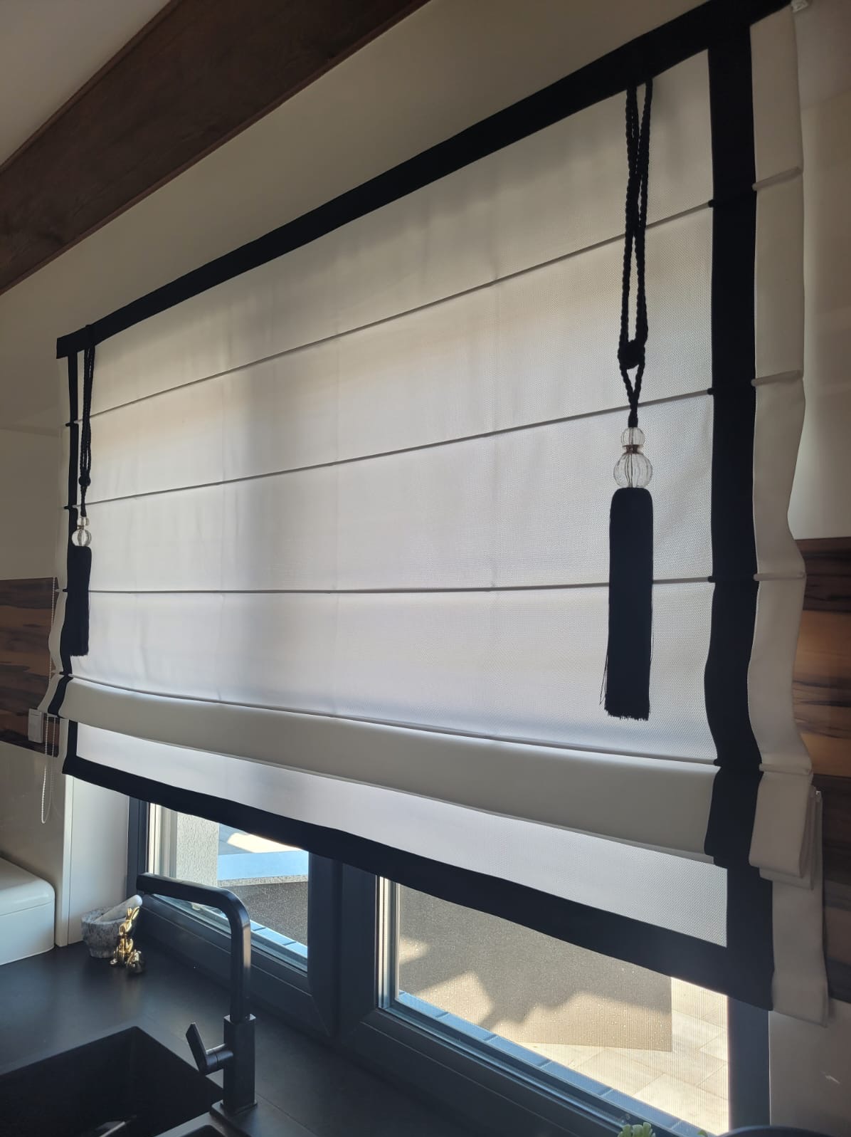 Elegancka, biała roleta rzymska z czarnymi akcentami i ozdobnymi frędzlami, zamontowana w oknie nad czarnym zlewem kuchennym. Widoczne drewniane elementy wykończenia.