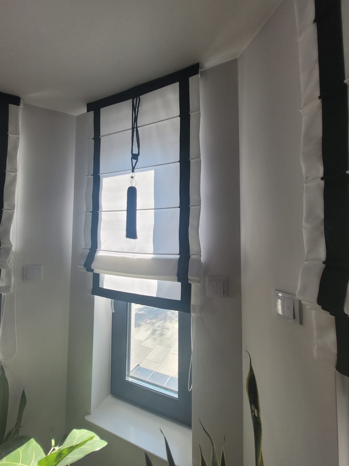 Elegancka roleta rzymska w kolorze białym z czarnymi akcentami, zamontowana w oknie. Dekoracyjna zawieszka dodaje uroku. Widok na zewnątrz przez okno.
