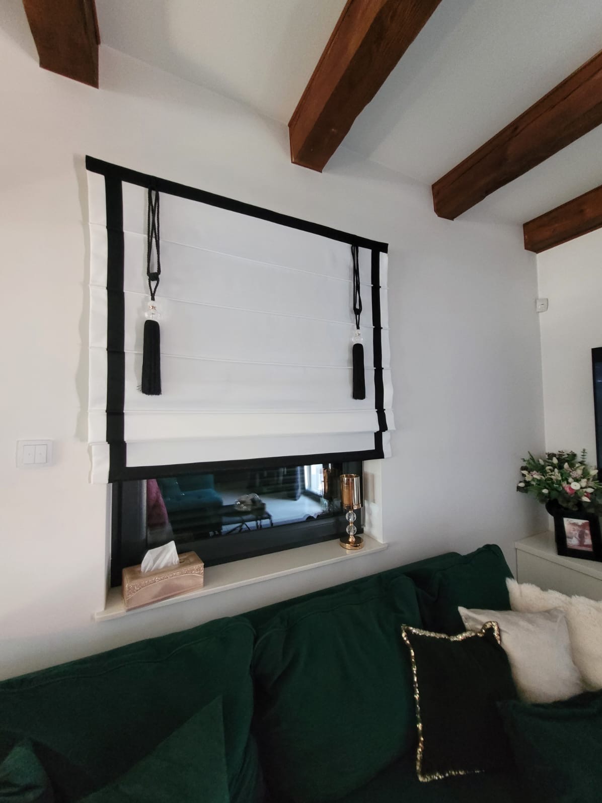 Elegancka roleta rzymska w kolorze białym z czarnymi akcentami, zwisającymi ozdobnymi chwostami, zamontowana w oknie. Widoczne drewniane belki stropowe.