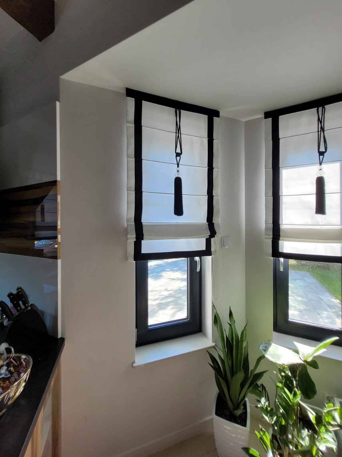 Dwie rolety rzymskie w czarno-białej kolorystyce, z dekoracyjnymi frędzlami, zamontowane w oknach z czarnymi ramami, widok z bliska, z roślinami doniczkowymi w tle.