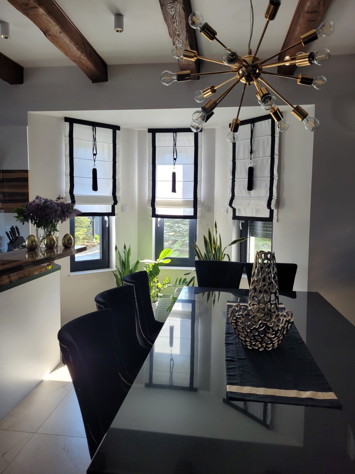 Eleganckie rolety rzymskie w czarno-białej kolorystyce, zdobiące okna jadalni z nowoczesnym żyrandolem i ciemnymi krzesłami przy szklanym stole.