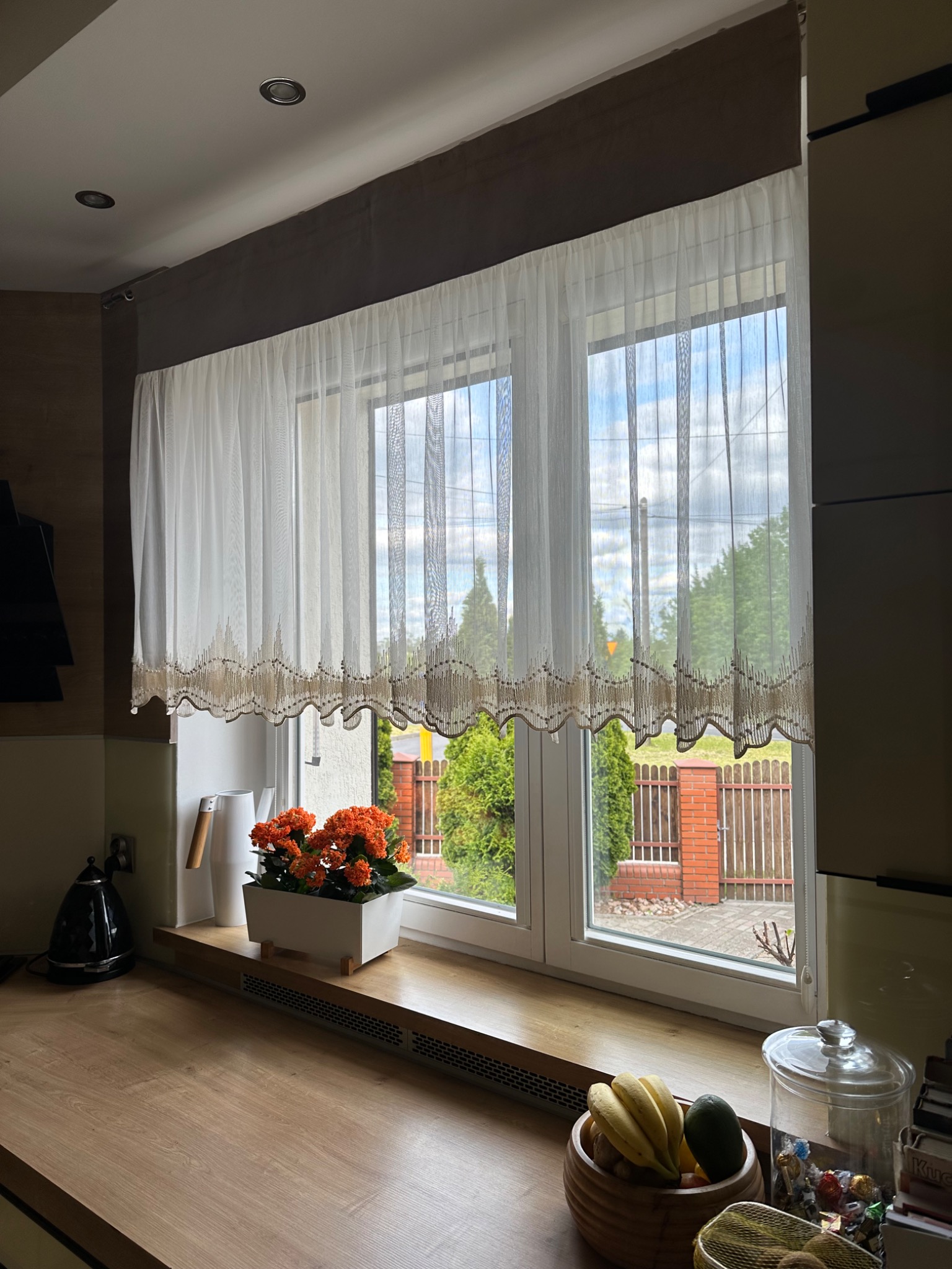 Delikatna, biała zasłona z ozdobnym, brązowym wykończeniem w oknie kuchni z widokiem na ogród. Na parapecie kwiaty i dekoracyjne przedmioty.
