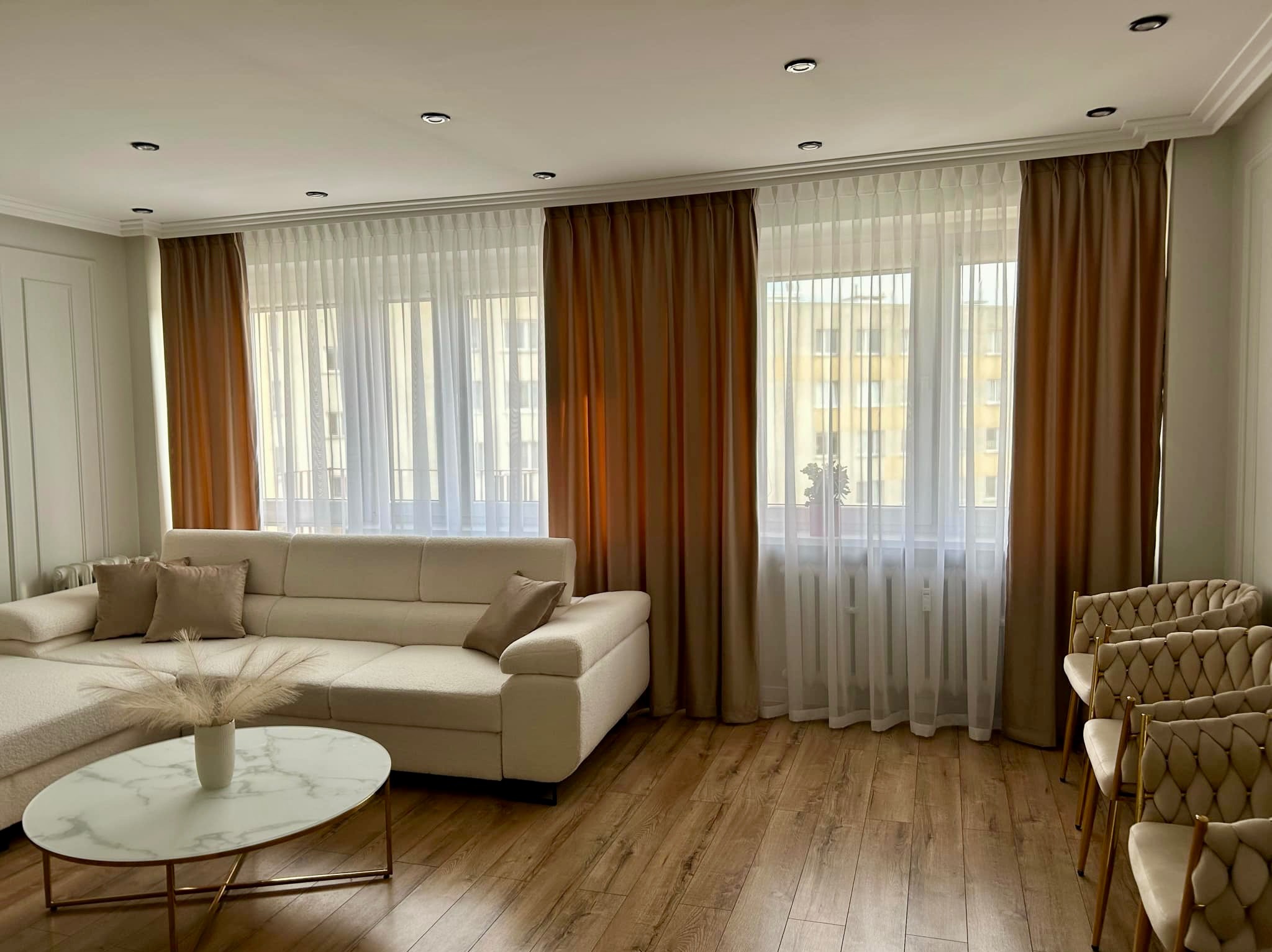 Salon z beżową sofą i marmurowym stolikiem. Okna przesłonięte brązowymi zasłonami i białymi firanami. Podłoga wyłożona panelami w odcieniach brązu.