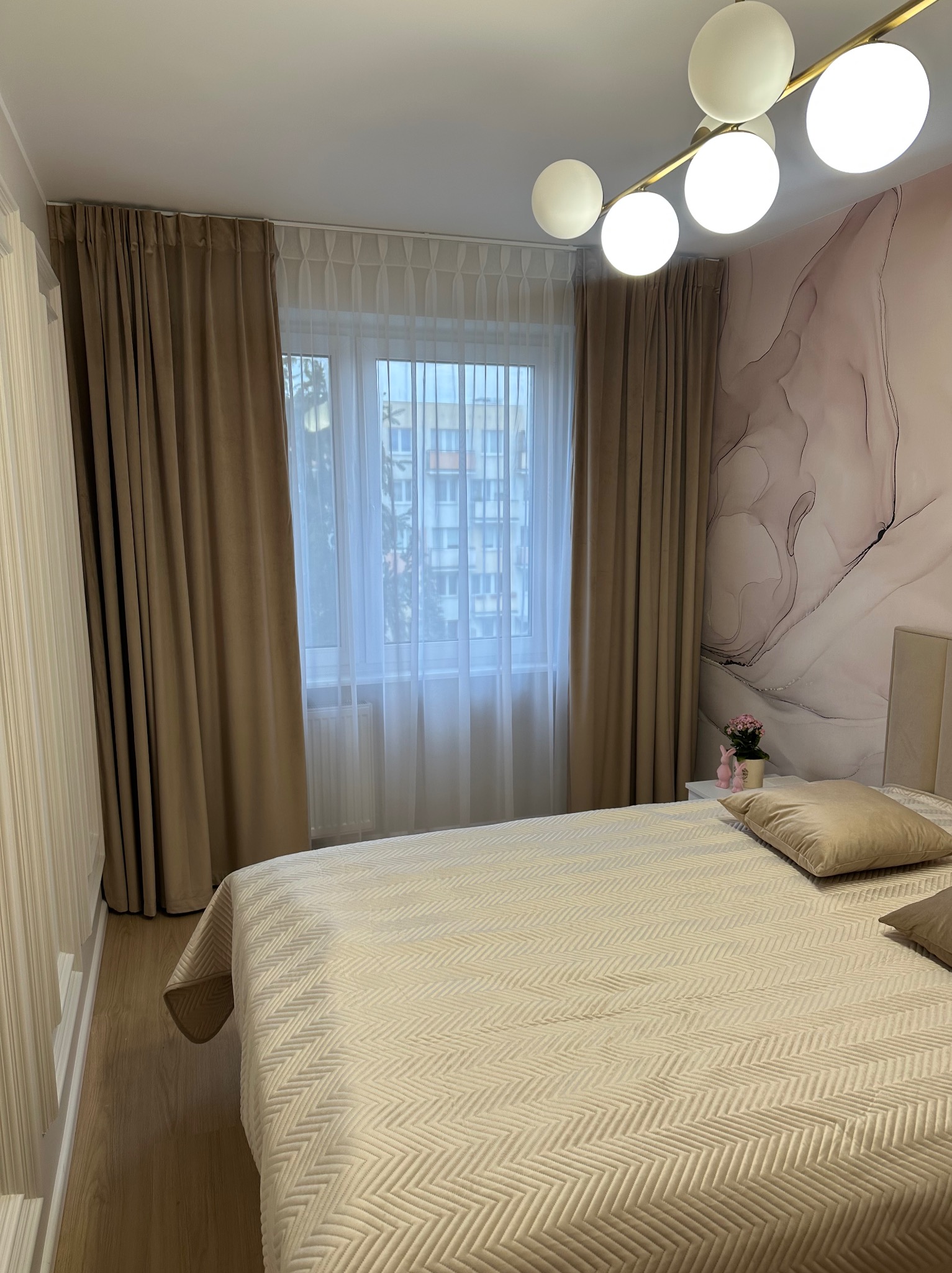 Beżowe zasłony zaciemniające w sypialni z wzorzystą tapetą i dekoracyjną lampą. Widok na okno z białymi firanami i budynkiem w tle.