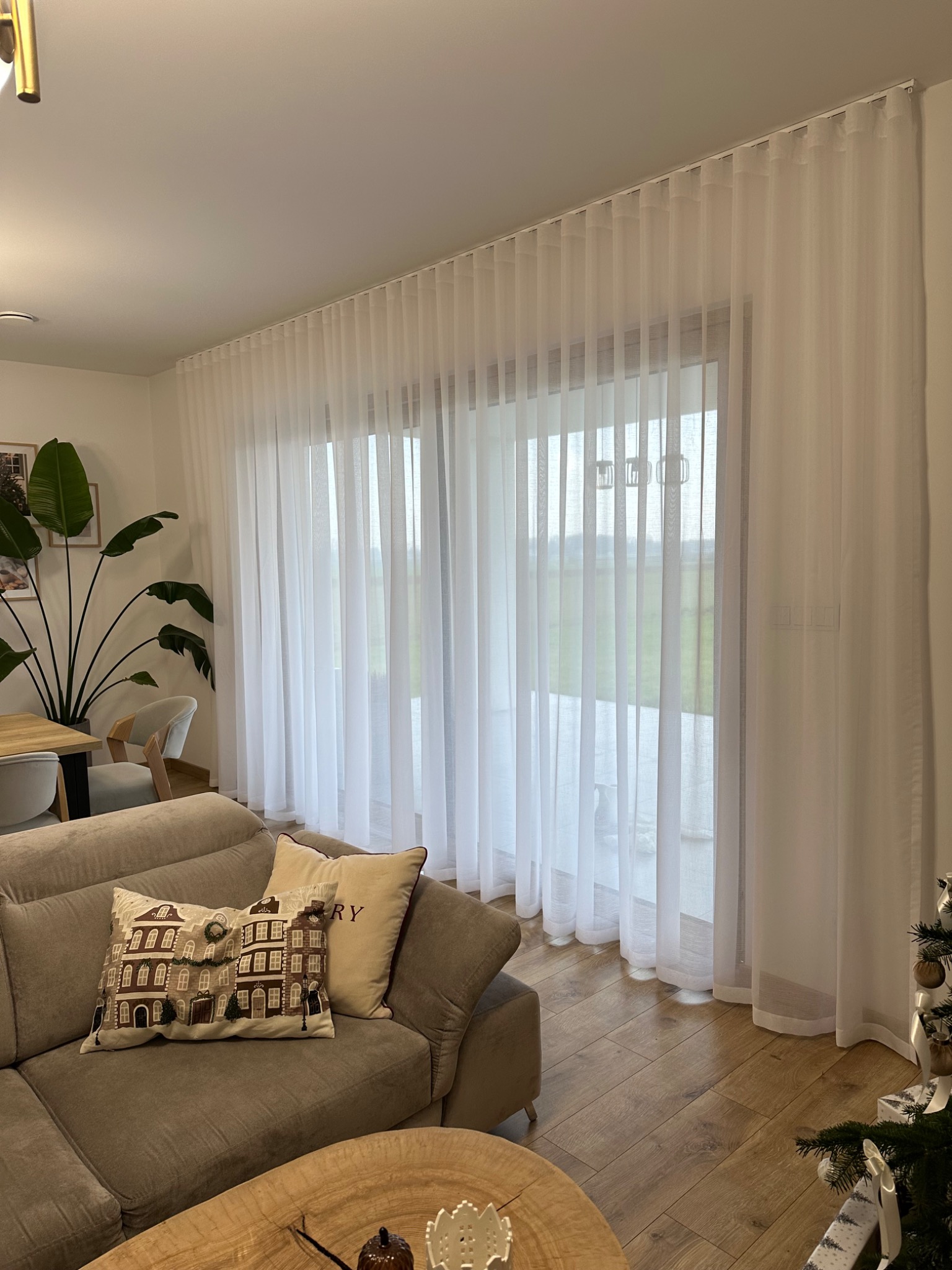 Jasne, zwiewne zasłony woalowe w salonie z drewnianą podłogą i beżową sofą. Dekoracyjne poduszki i roślina doniczkowa dodają przytulności wnętrzu.