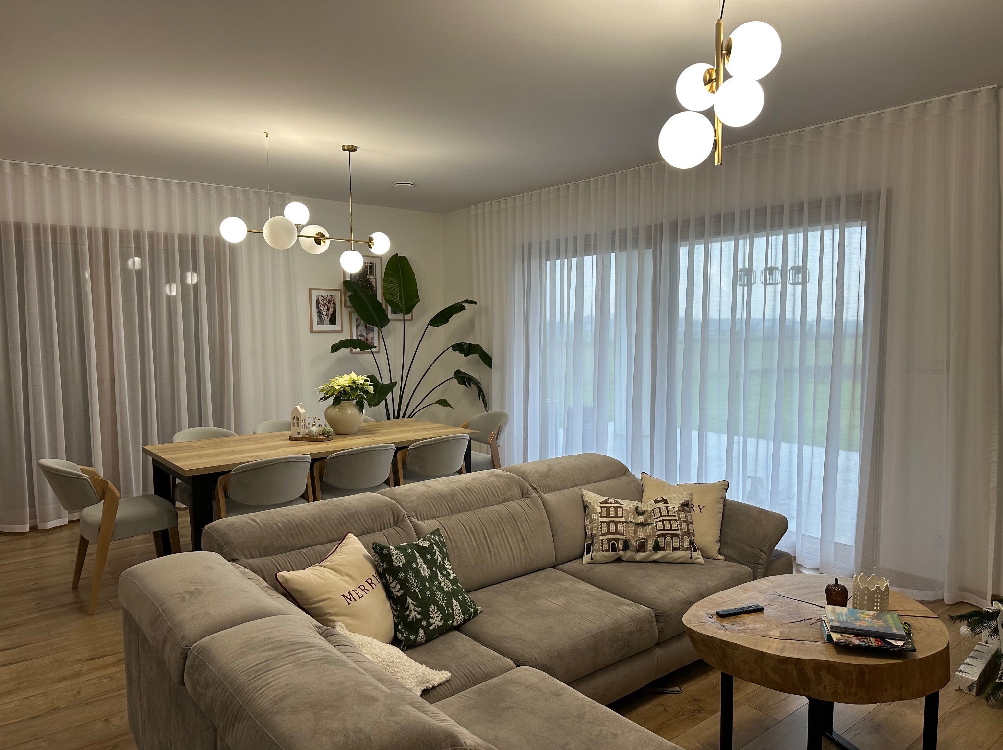 Jasny salon z beżową sofą i drewnianym stołem. Okno zasłonięte długimi, białymi, zwiewnymi zasłonami. Nowoczesne lampy wiszące.