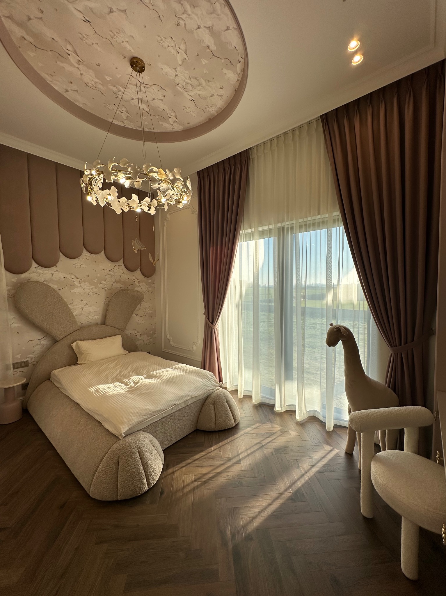 Pokój dziecięcy z zasłonami w odcieniach brązu i beżu, wpadające promienie słońca, pluszowy koń przy oknie, łóżko w kształcie królika, ozdobny żyrandol i malowidło na suficie.