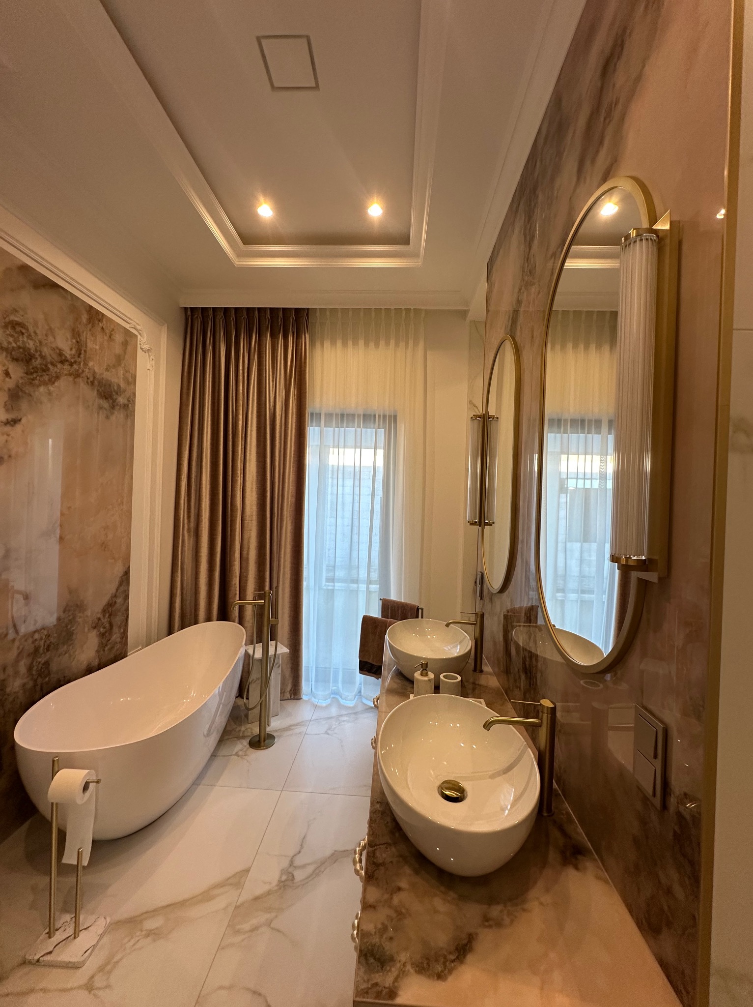 Elegancka łazienka z marmurowymi ścianami, owalną wanną i złotymi akcentami. Zasłony w odcieniach brązu wpuszczają delikatne światło. Lustra w złotych ramach.