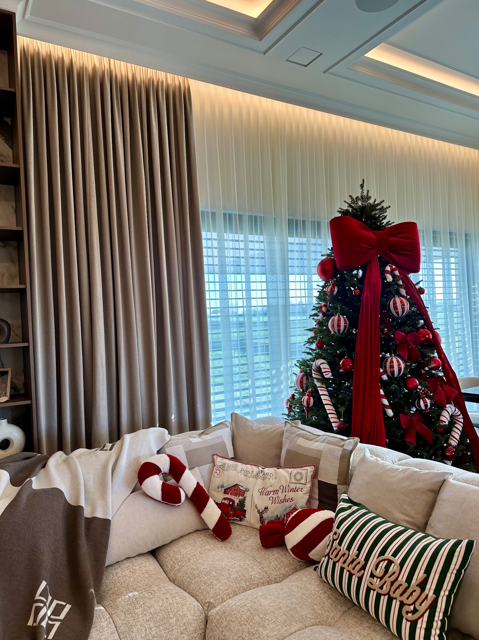Beżowe, grube zasłony w salonie z choinką, dekoracjami świątecznymi i kanapą z poduszkami. Widoczne światło dzienne wpadające przez okno.