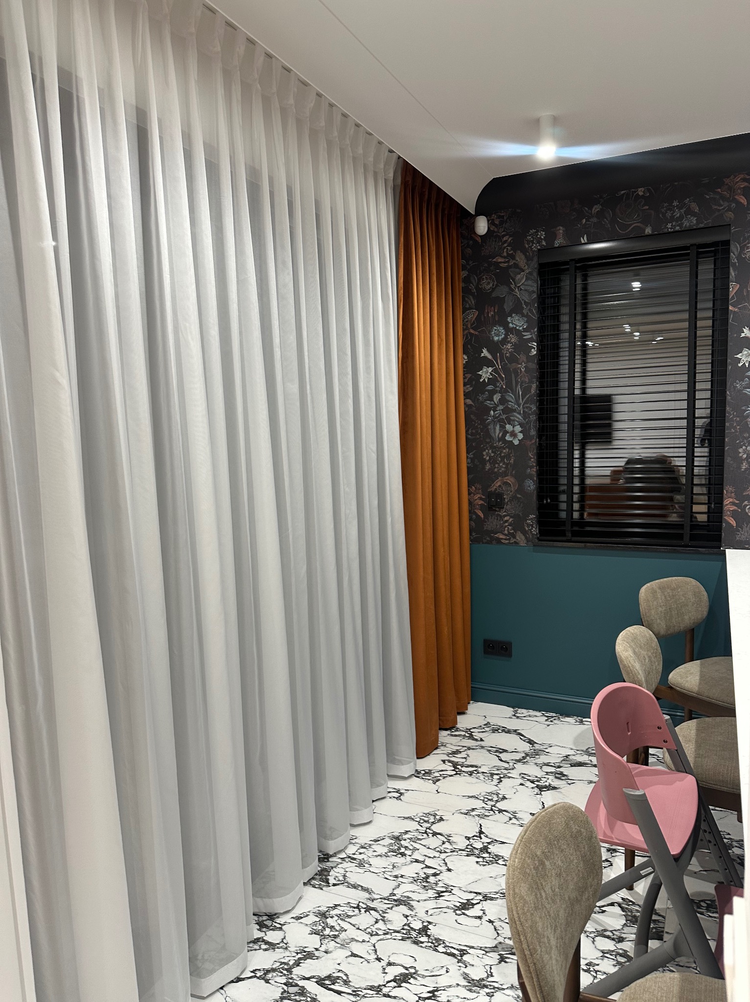 Delikatne, białe zasłony woalowe w nowoczesnym wnętrzu z marmurową podłogą i akcentami w kolorze musztardowym. Widoczne krzesła i okno z żaluzjami.