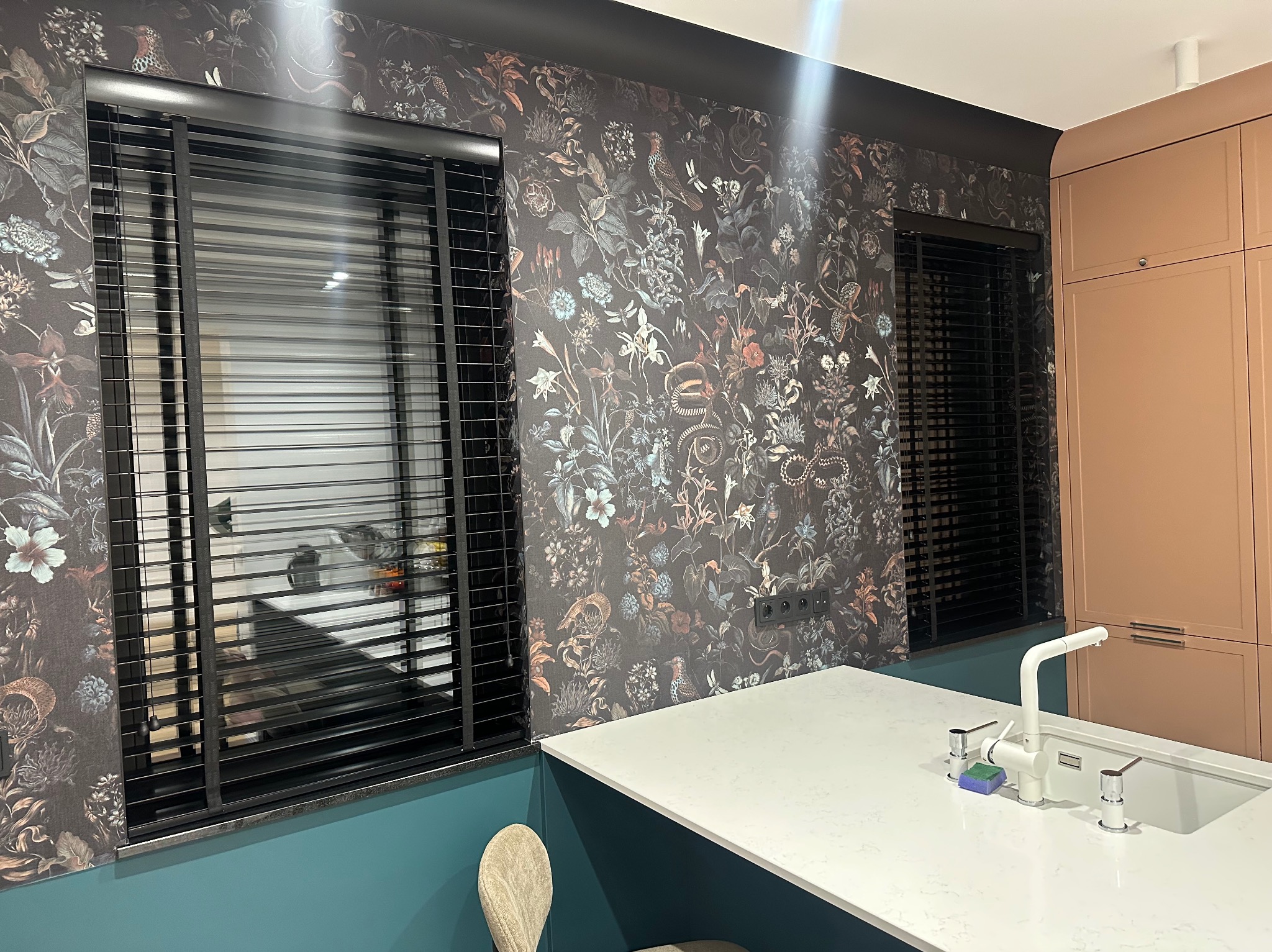 Czarne żaluzje poziome w oknach kuchni z tapetą w kwiaty i wzory. Blat kuchenny z białego kamienia i bateria. Nowoczesne wykończenie wnętrza.