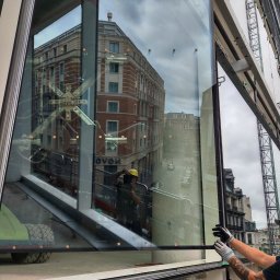 BKM Bud Katarzyna Wolińska - Montaż dużej szyby okiennej z widocznym odbiciem budynku w tafli szkła. Pracownik w rękawicach stabilizuje szybę na tle konstrukcji rusztowania.