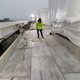 BKM Bud Katarzyna Wolińska - Pracownik w odblaskowej kurtce łączy płyty Aquapanel na zewnątrz budynku, używając palnika. Widoczne narzędzia i materiały budowlane. Szare niebo w tle.