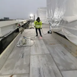 Pracownik w odblaskowej kurtce łączy płyty Aquapanel na zewnątrz budynku, używając palnika. Widoczne narzędzia i materiały budowlane. Szare niebo w tle.