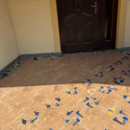 Układanie brązowych płytek na zewnątrz budynku z użyciem klipsów poziomujących. Widoczne drzwi wejściowe i fragment ściany. Etap prac glazurniczych.