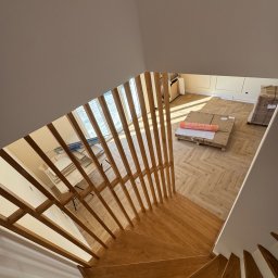 BKM Bud Katarzyna Wolińska - Drewniane schody spiralne z ażurową balustradą, widok z góry na parkiet w jodełkę i materiały budowlane. Jasne wnętrze w trakcie wykańczania.