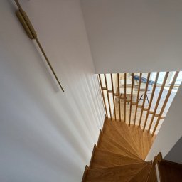 BKM Bud Katarzyna Wolińska - Drewniane schody spiralne z balustradą z pionowych listew, widok z góry, białe ściany i złoty kinkiet. W tle widoczne narzędzia i materiały budowlane.
