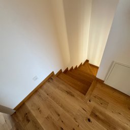 BKM Bud Katarzyna Wolińska - Drewniane schody z parkietem w jasnym wnętrzu. Widoczne stopnie i podłoga z naturalnego drewna, białe ściany z gniazdkiem elektrycznym. Ujęcie z góry.