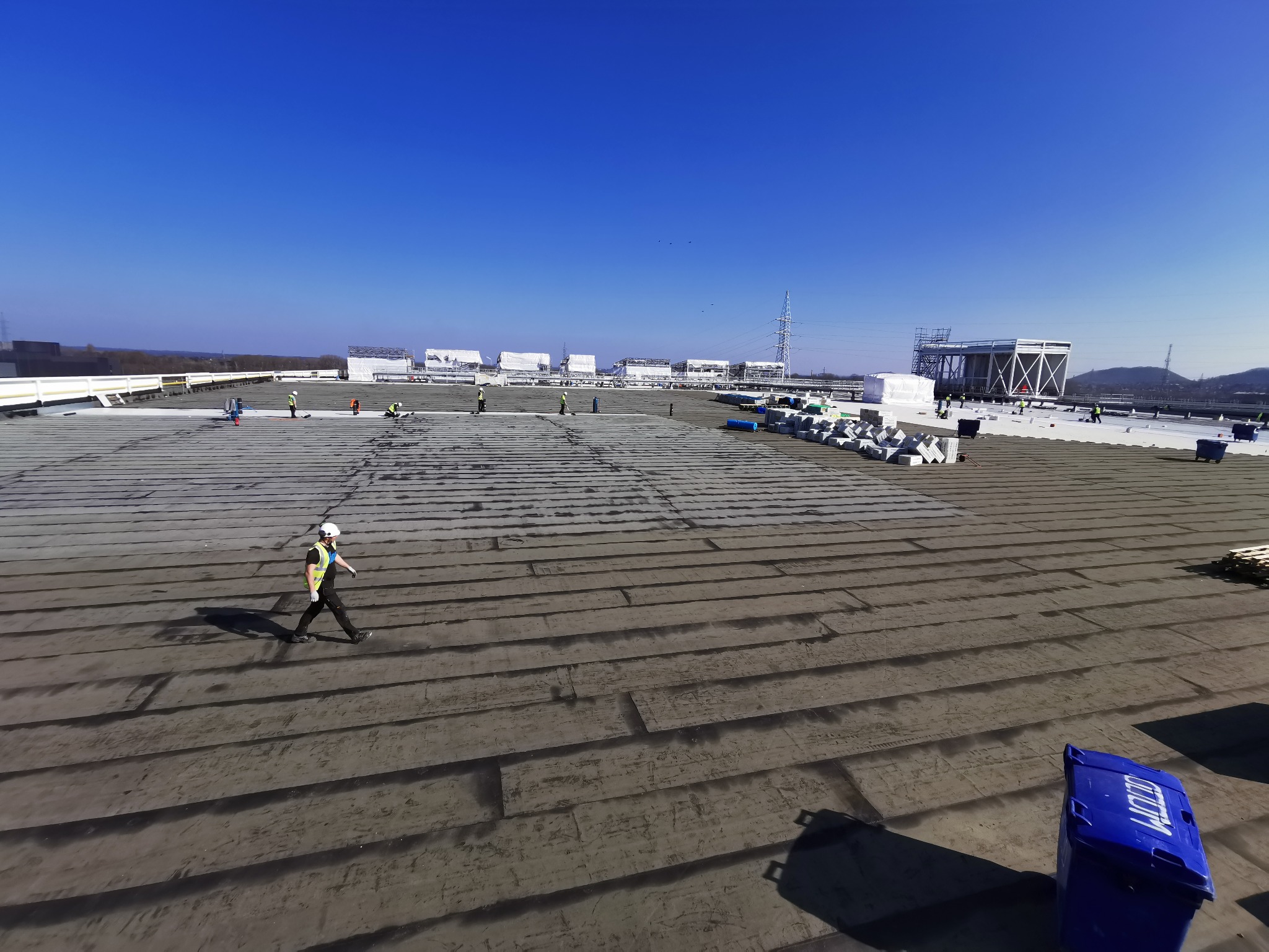 Rozległy dach pokryty papą, na którym pracują robotnicy w kaskach. Widoczne materiały budowlane i elementy konstrukcyjne. Jasne niebo w tle.