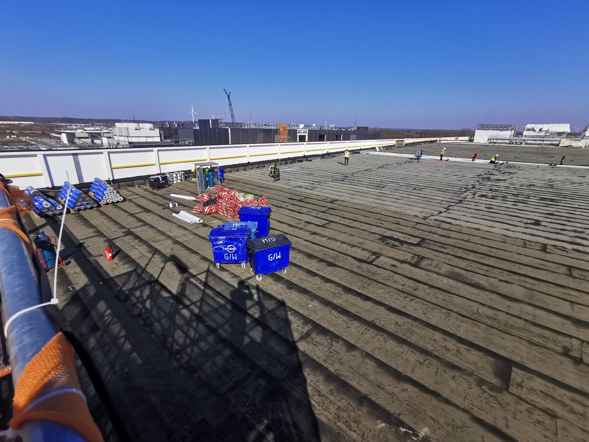 Prace na dachu budynku przemysłowego: ekipa w kaskach, materiały budowlane i narzędzia. Widok z góry, słoneczny dzień. W tle zabudowania i dźwig.