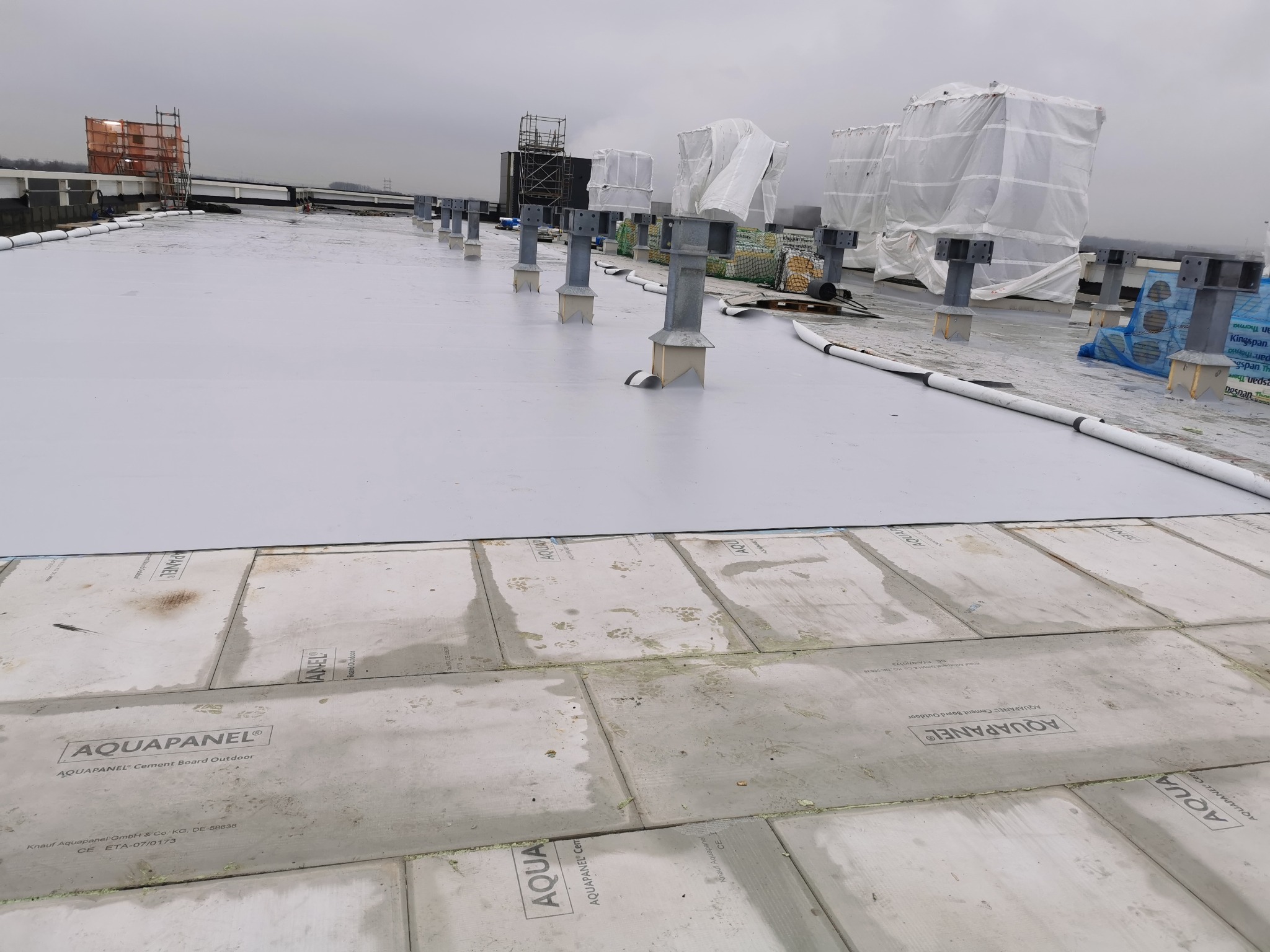 Montaż płyt Aquapanel na dachu budynku. Widoczne płyty cementowe, metalowe wsporniki i biała membrana. W tle rusztowania i zabudowy zabezpieczone folią.