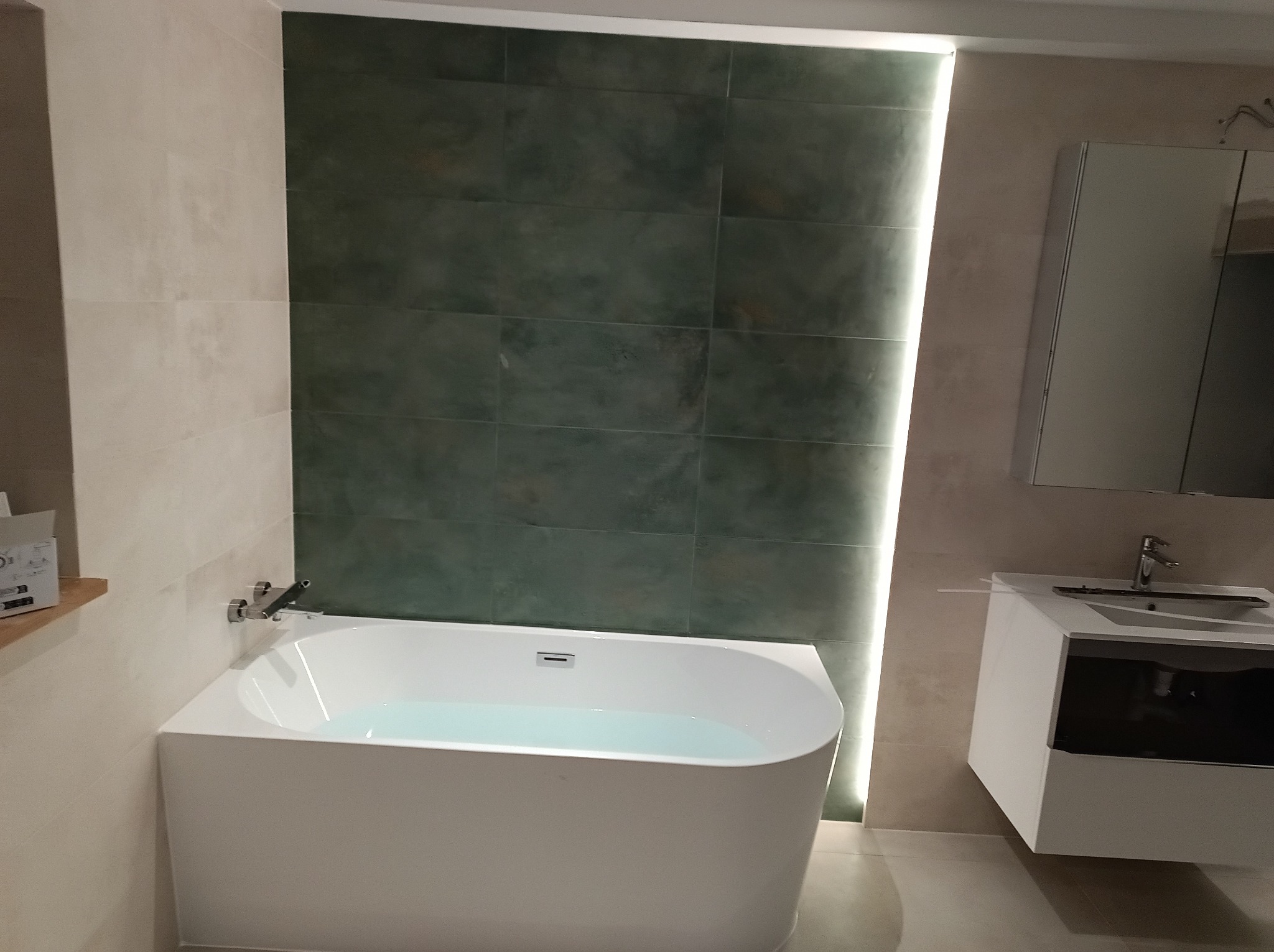 Nowoczesna łazienka z wanną wypełnioną wodą, umywalką z szafką i lustrem. Ściany wyłożone płytkami w odcieniach beżu i zieleni. Oświetlenie LED podkreśla pionowy układ płytek.