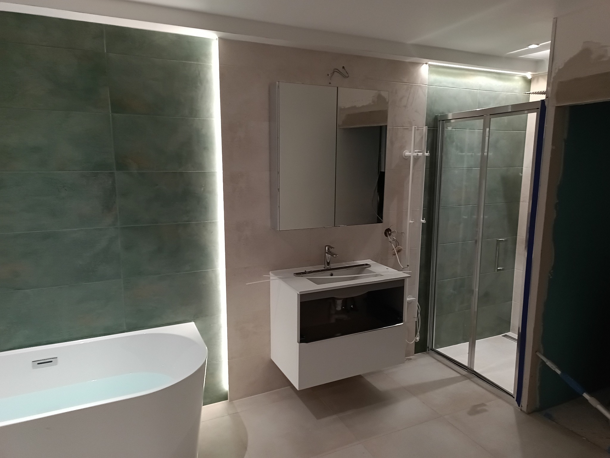 Nowoczesna łazienka z wanną, umywalką z szafką i kabiną prysznicową. Ściany wyłożone płytkami w odcieniach zieleni i beżu, oświetlenie LED. Minimalistyczny design.