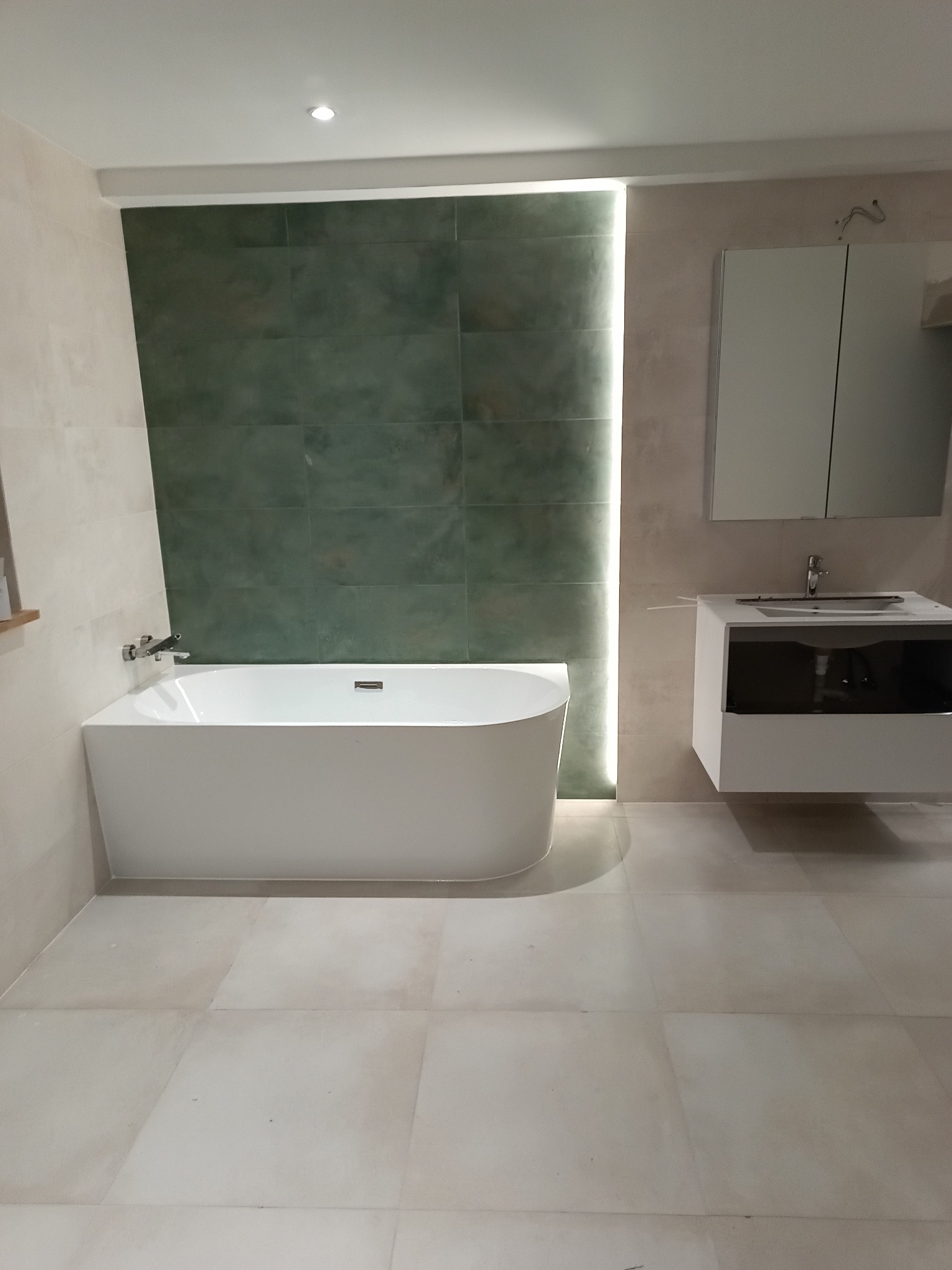 Nowoczesna łazienka z wolnostojącą wanną, zieloną ścianą z płytek i podwieszaną szafką z umywalką. Jasne płytki podłogowe i subtelne oświetlenie.