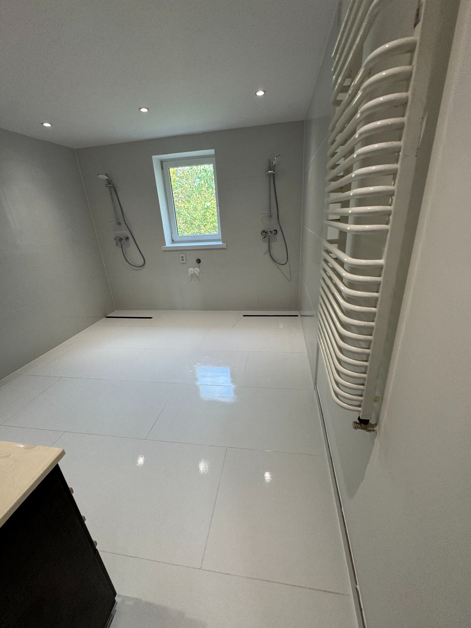 Wykończona łazienka w szarościach z białymi płytkami, grzejnikiem drabinkowym i dwoma prysznicami. Okno z widokiem na zieleń. Minimalistyczny design.