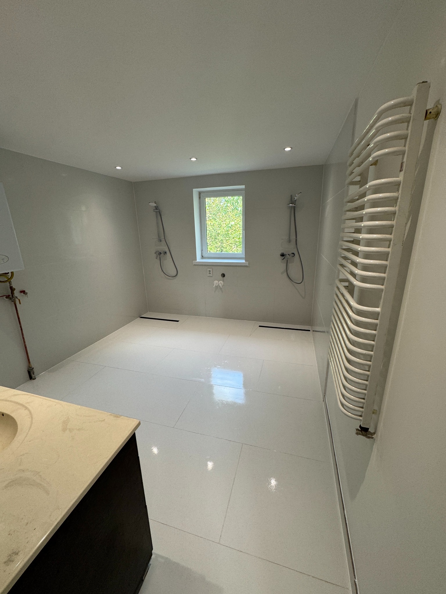 Jasna łazienka po remoncie z białymi płytkami, grzejnikiem drabinkowym i dwoma prysznicami. Widoczne okno z zielenią na zewnątrz i fragment szafki z umywalką.