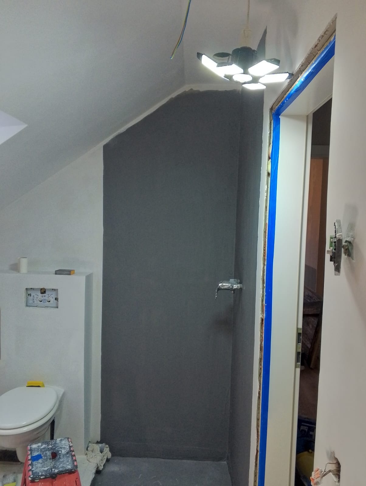 Szara, wykończona strefa prysznicowa w łazience ze skośnym sufitem, widoczna toaleta i przygotowane do montażu drzwi z niebieską taśmą zabezpieczającą.