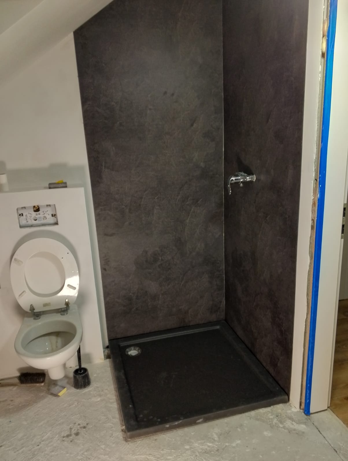 Wykończona kabina prysznicowa z ciemnymi płytami imitującymi kamień, widoczny brodzik i bateria. W tle toaleta i fragment ściany z instalacją.