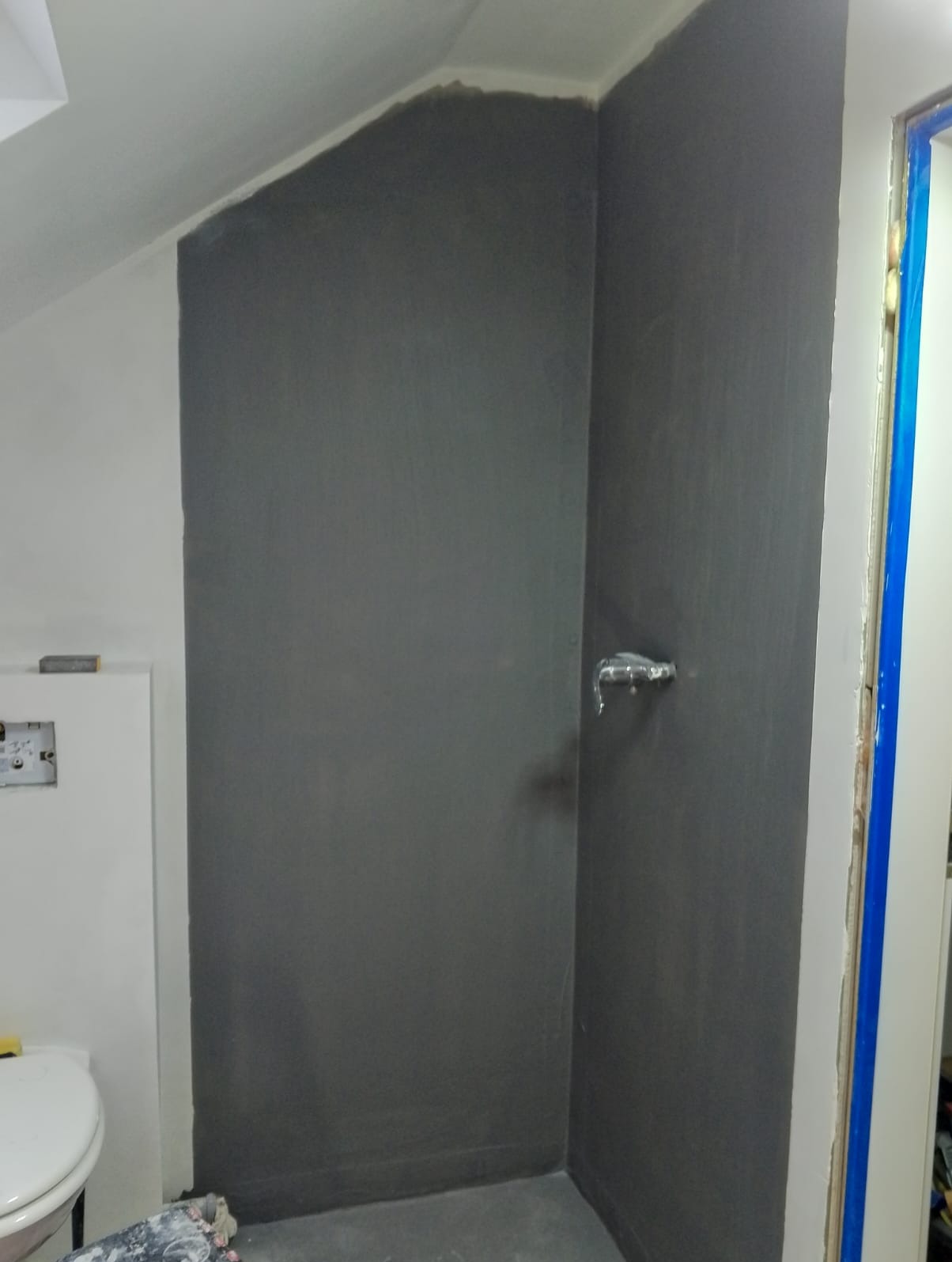 Ściany w szarości w trakcie prac wykończeniowych w łazience. Widoczny fragment toalety i zabezpieczone niebieską folią obramowanie drzwi.