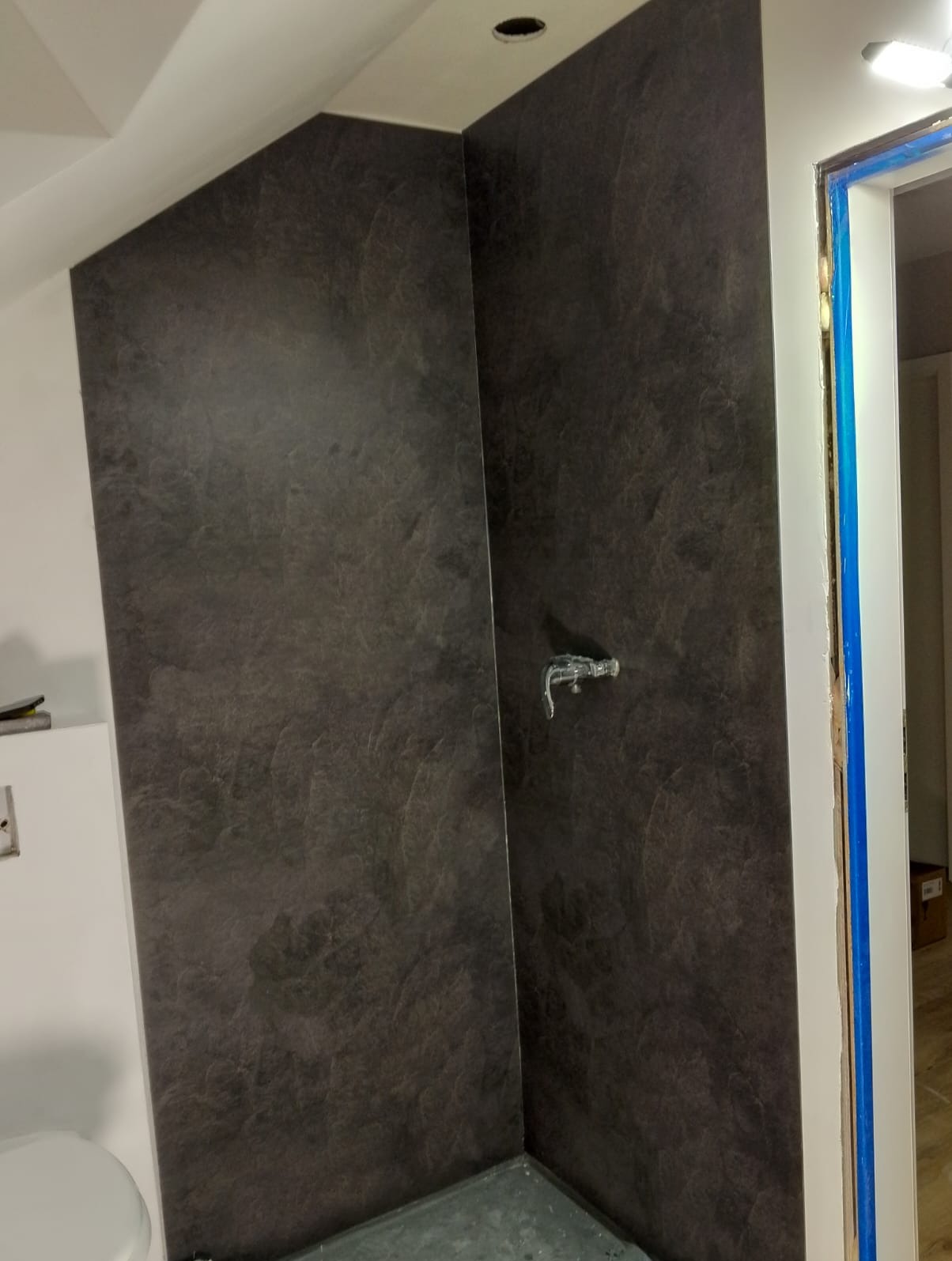 Wykończona ściana w łazience z ciemnymi płytkami imitującymi kamień, widoczna bateria prysznicowa i fragment futryny drzwiowej zabezpieczonej folią.