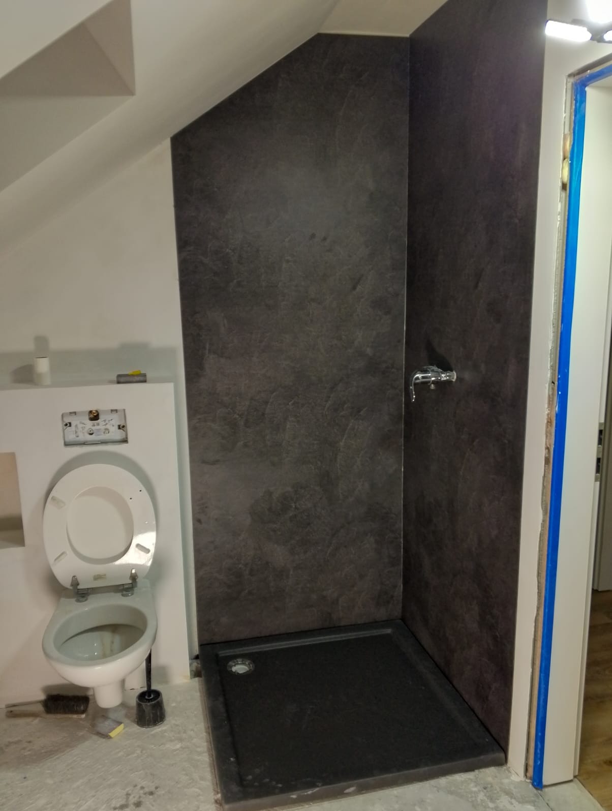 Szara kabina prysznicowa z brodzikiem i toaleta w trakcie montażu w łazience na poddaszu. Widoczne narzędzia i niedokończone wykończenie.