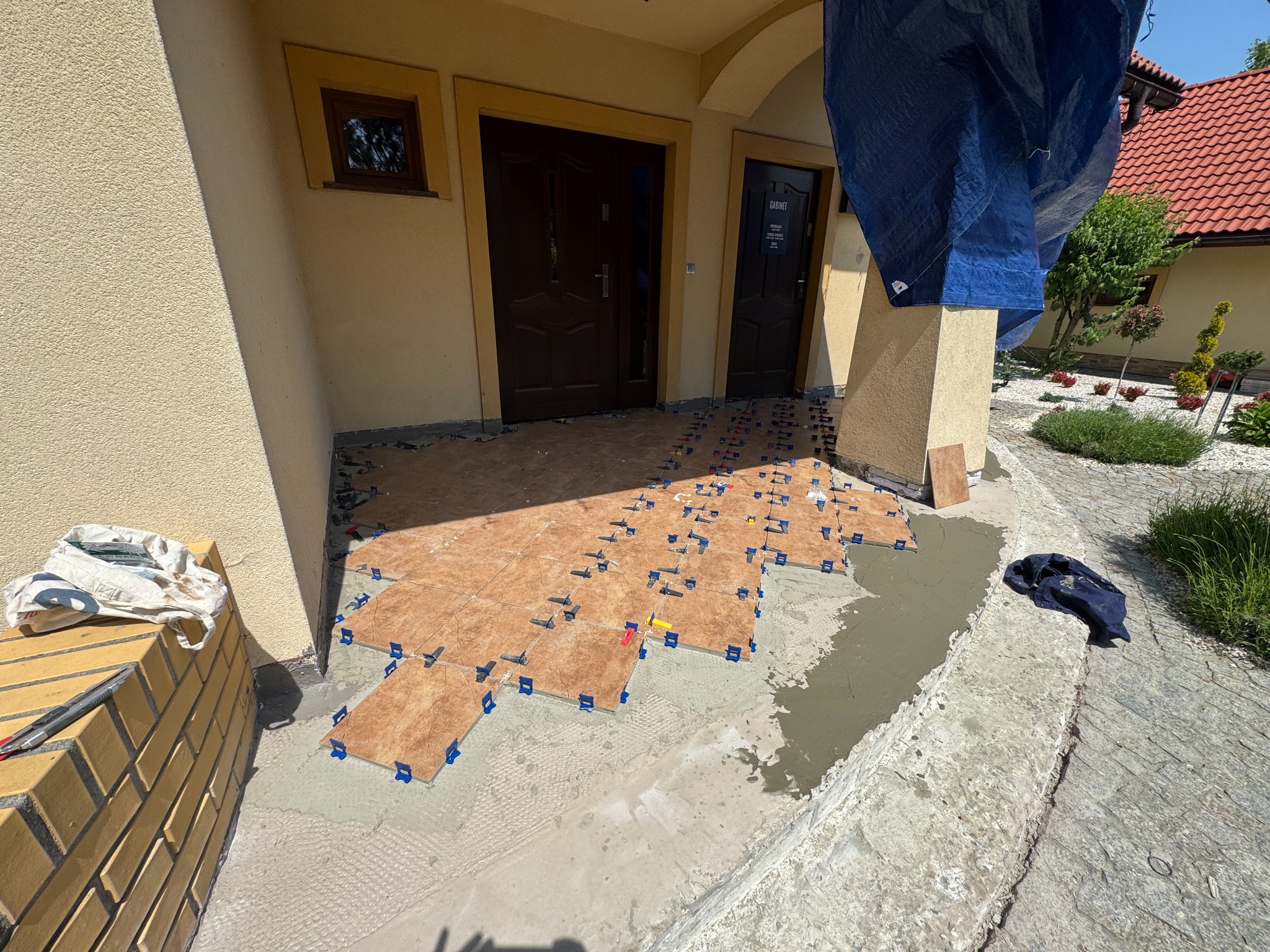 Układanie brązowych płytek na zewnątrz budynku z użyciem klipsów poziomujących. Widoczne narzędzia glazurnicze i fragment elewacji. Prace w toku, etap układania płytek.