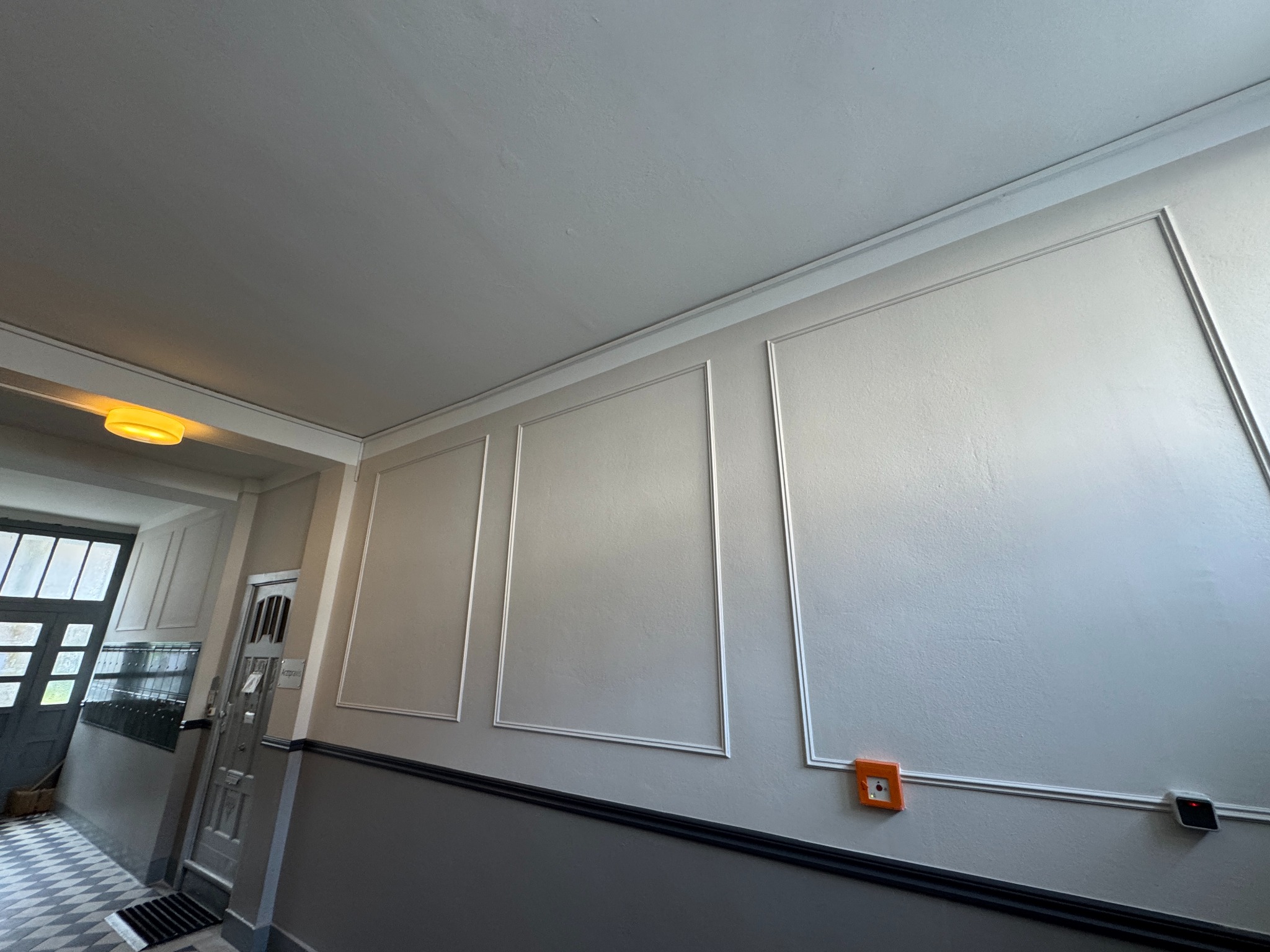 Wykończona klatka schodowa z panelami ściennymi w odcieniach szarości i bieli. Widoczne drzwi wejściowe i fragment podłogi w szachownicę. Oświetlenie sufitowe.