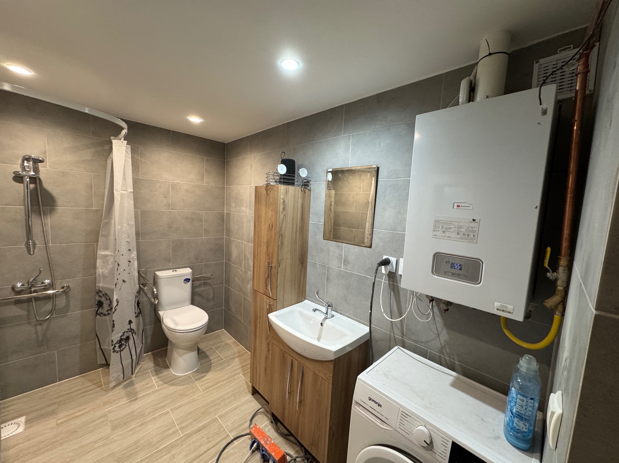 Wykończona łazienka w szarościach z kabiną prysznicową, umywalką z szafką, pralką i piecem gazowym. Widoczne płytki na ścianach i podłodze, oświetlenie sufitowe.